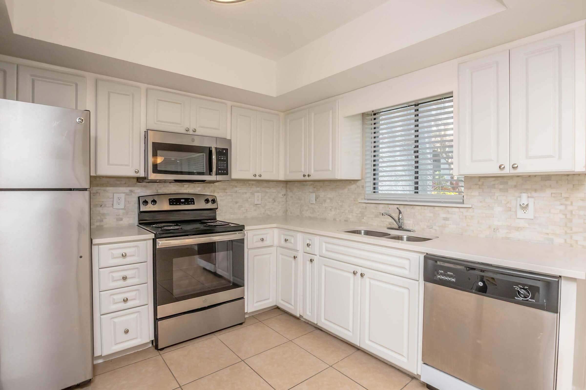 Modern kitchen featuring white cabinetry, stainless steel appliances including a refrigerator, microwave, and oven, and a double sink. The space has beige tile flooring and a window with blinds, allowing natural light to illuminate the area.