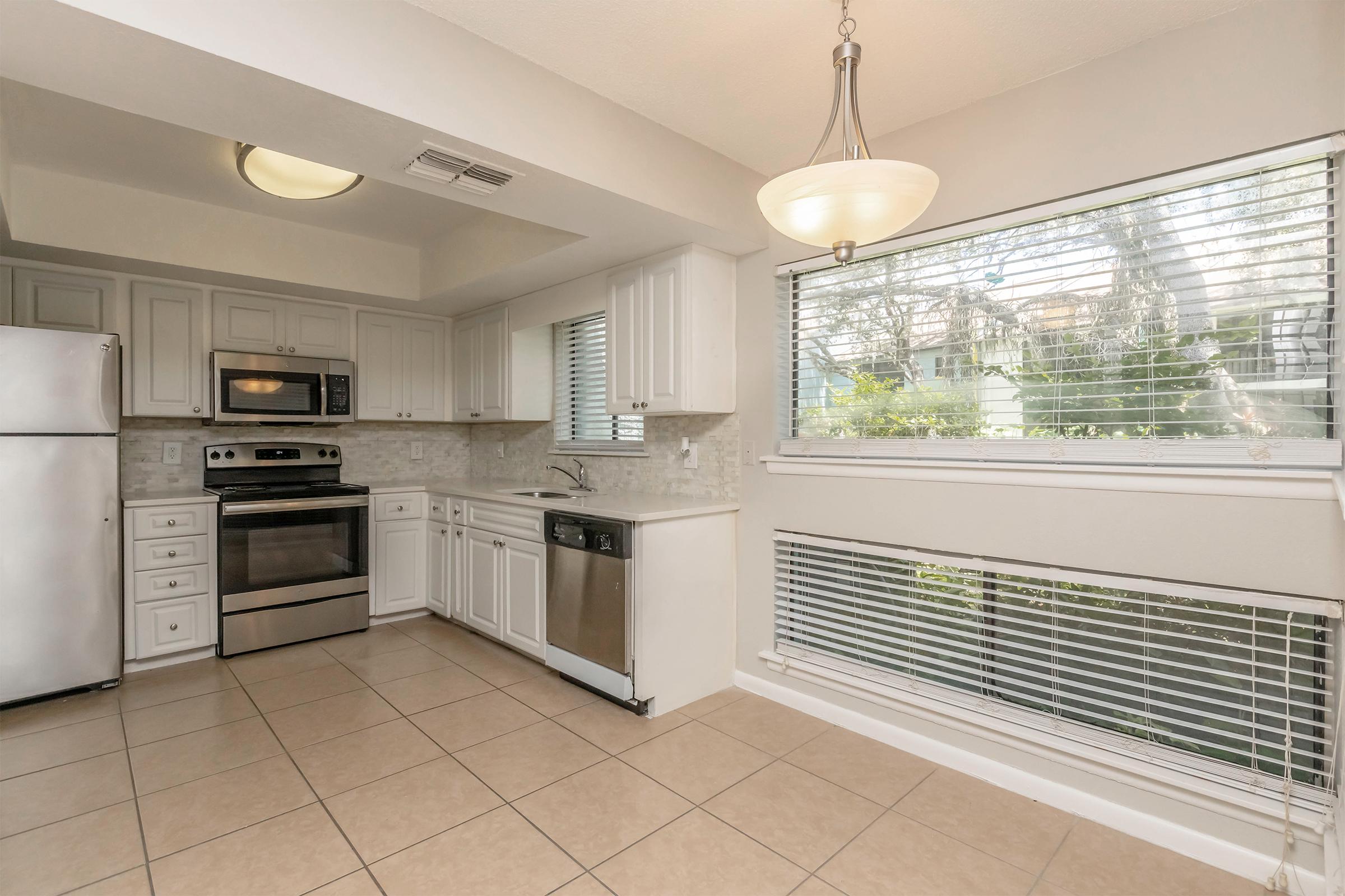 Bright and modern kitchen featuring white cabinetry, stainless steel appliances including a refrigerator, microwave, and oven. The space has a tiled floor and large windows with blinds, providing natural light. A pendant light hangs over the dining area, creating an inviting atmosphere.