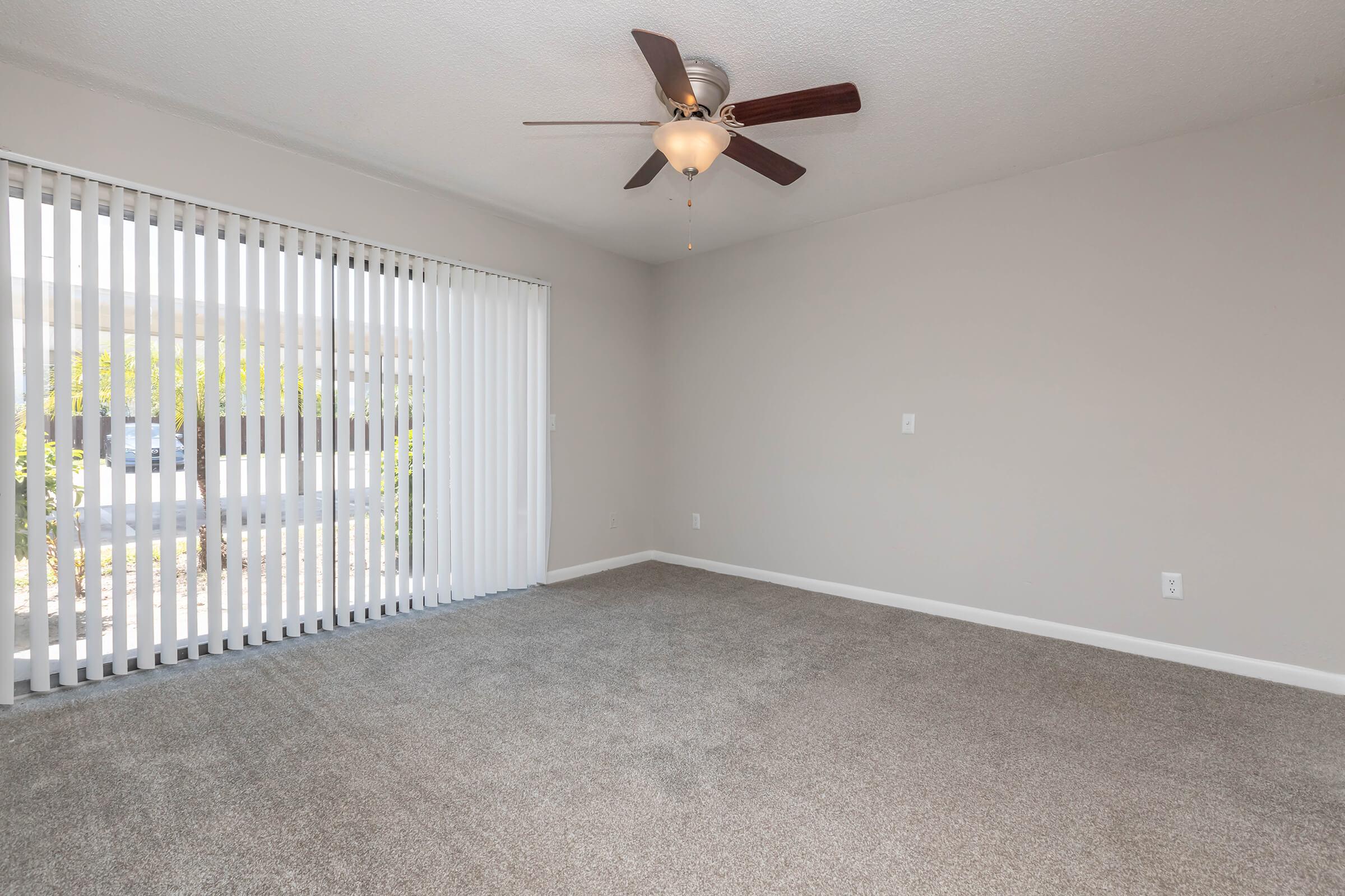 A spacious, empty room with light brown carpet and neutral-colored walls. It features a ceiling fan with wooden blades and large vertical blinds covering a window that lets in natural light. The room has a clean, open feel, ready for furniture arrangement.