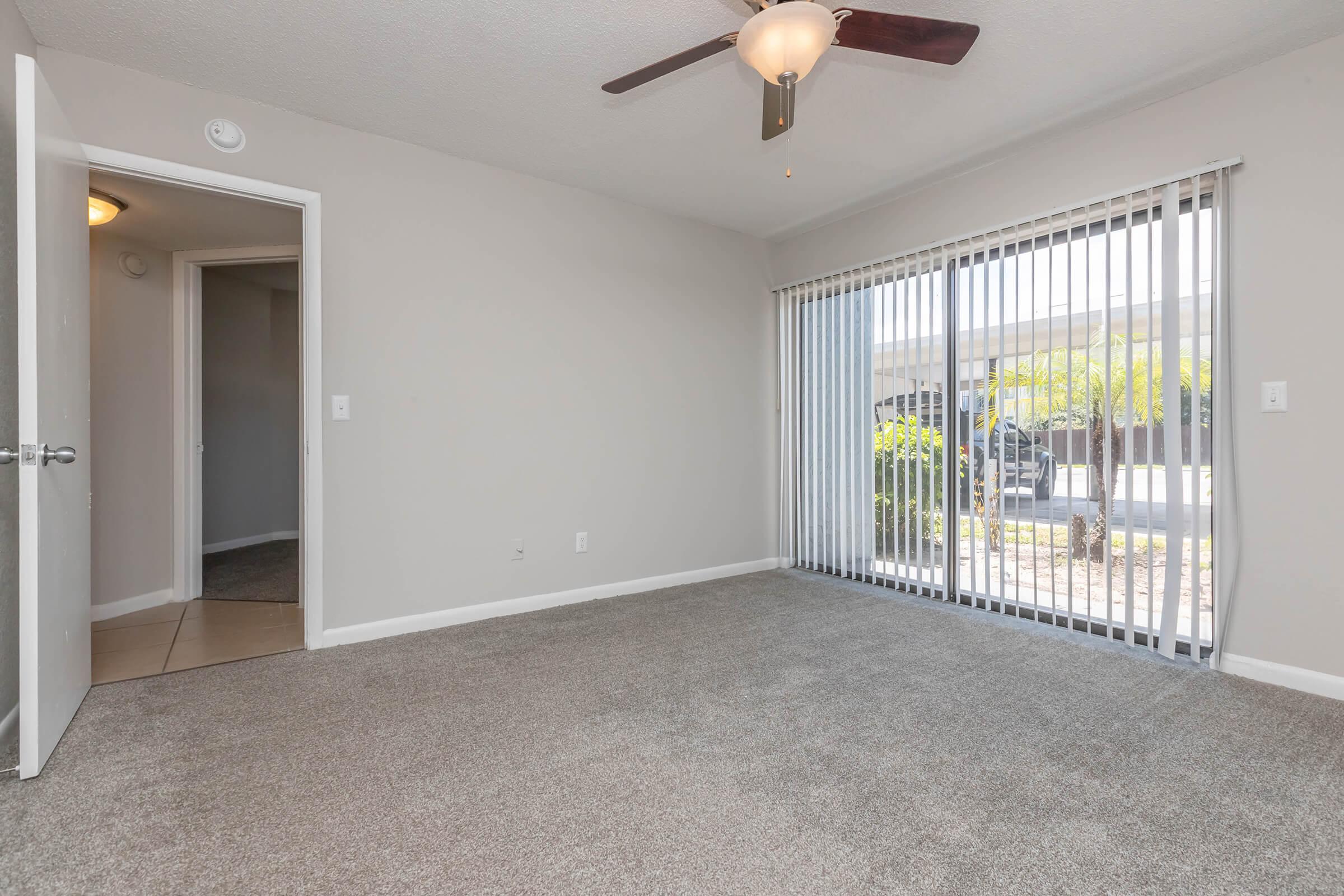 A well-lit, empty living room with carpet flooring, featuring a ceiling fan and large sliding glass doors that lead to an outdoor area. There is a doorway on the left leading to another room, and the walls are painted in a neutral color.