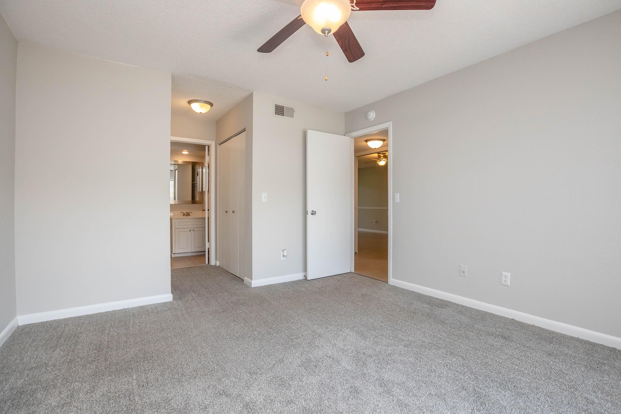 A spacious, light-filled bedroom featuring gray carpeting, a ceiling fan, and a neutral color scheme. The room has two doors leading to adjacent areas and a visible doorway to a bathroom, creating a sense of openness and accessibility.