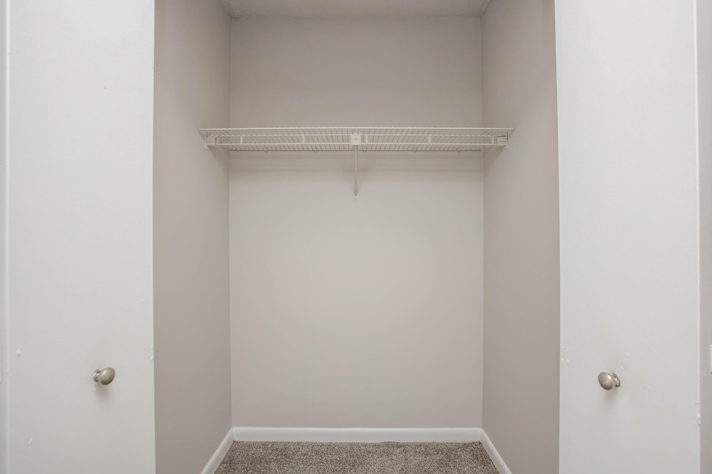 Empty closet with a single wire shelf mounted on the back wall. The walls are painted light grey, and the flooring is carpeted in a neutral tone. There are two doorknobs visible on the closet doors.