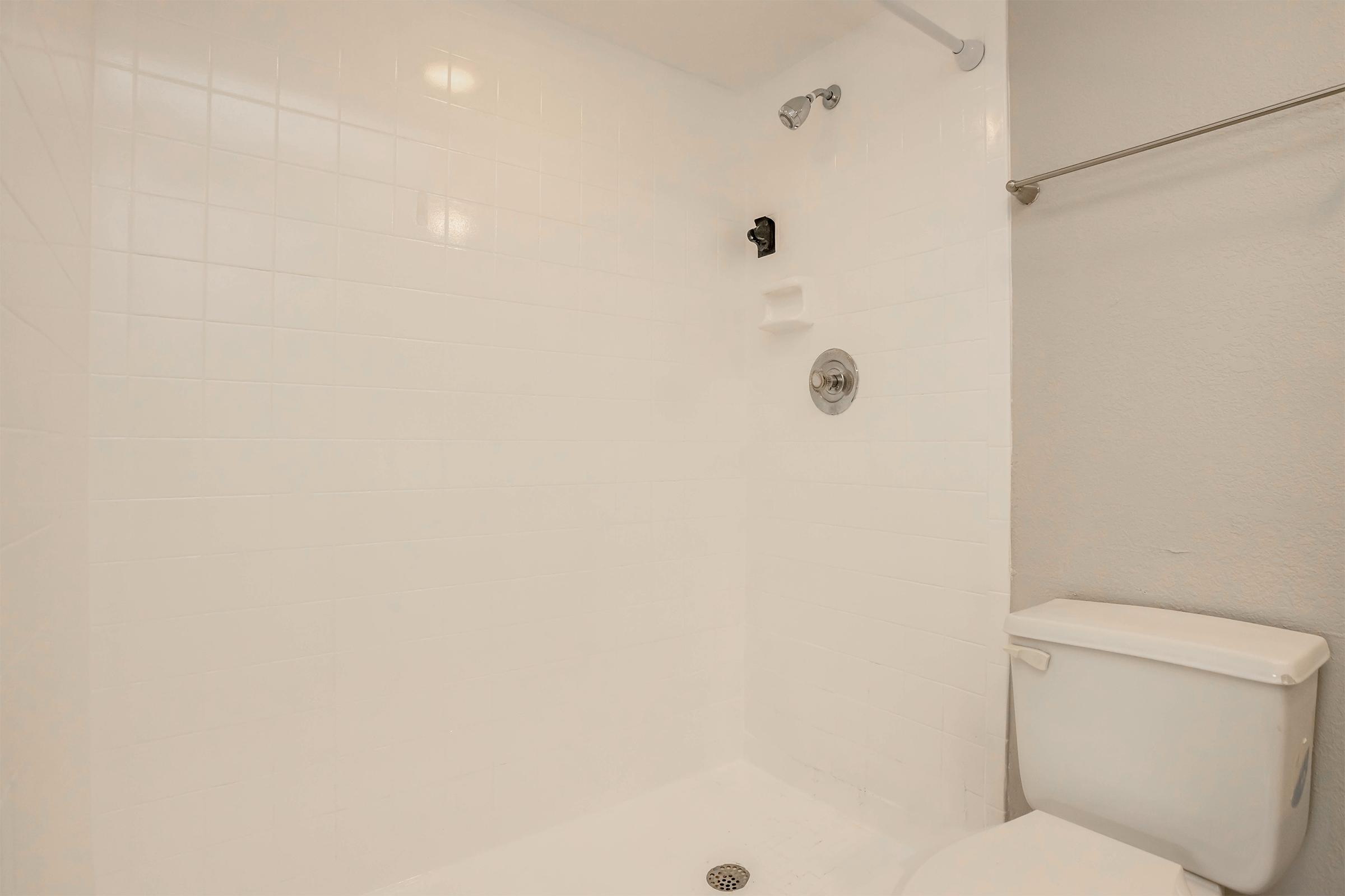 A bright, clean bathroom featuring a white tiled shower area with a showerhead, a wall-mounted soap holder, and a modern toilet. The space has neutral-colored walls and a simple, minimalist design.