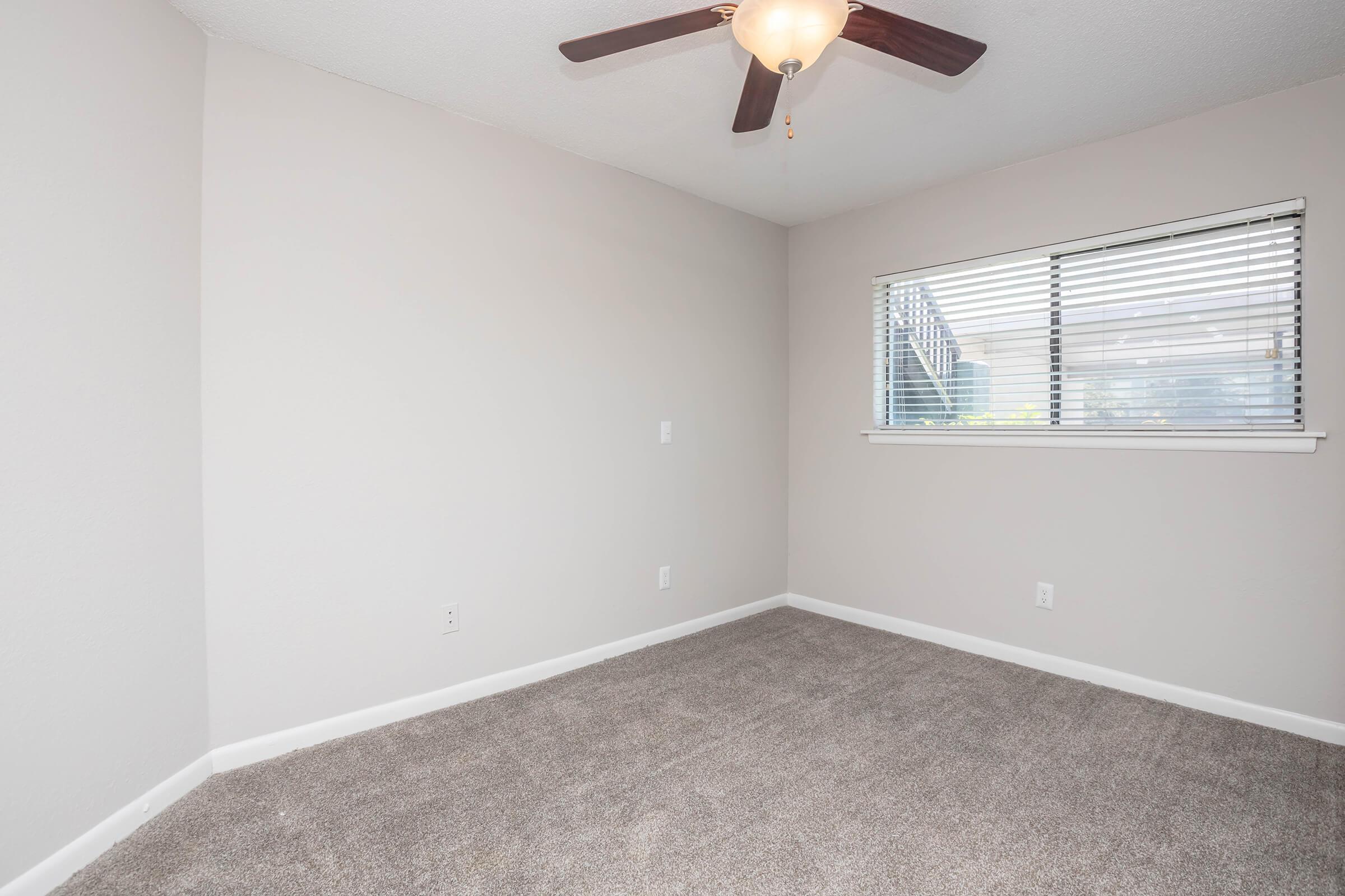A vacant room with light gray walls and carpeting. A ceiling fan with a light fixture is installed in the center. The room features a window with blinds, allowing natural light to enter. The space is uncluttered, creating a simple and clean appearance.