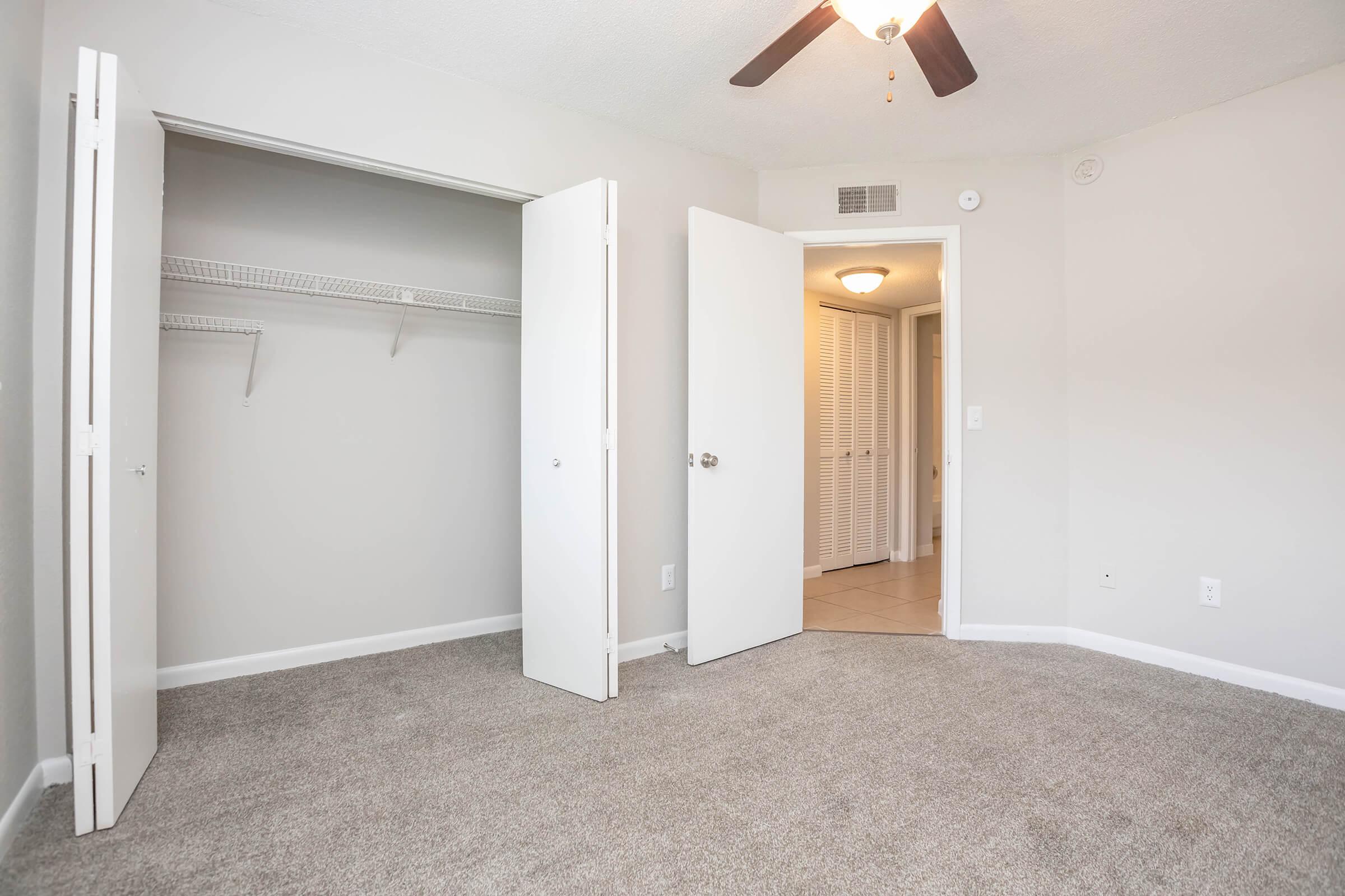 Spacious bedroom featuring light gray walls, a ceiling fan, and plush carpet. The room includes a double closet with open doors and a doorway leading to a hallway with additional storage. Natural light is present, enhancing the airy feel of the space.