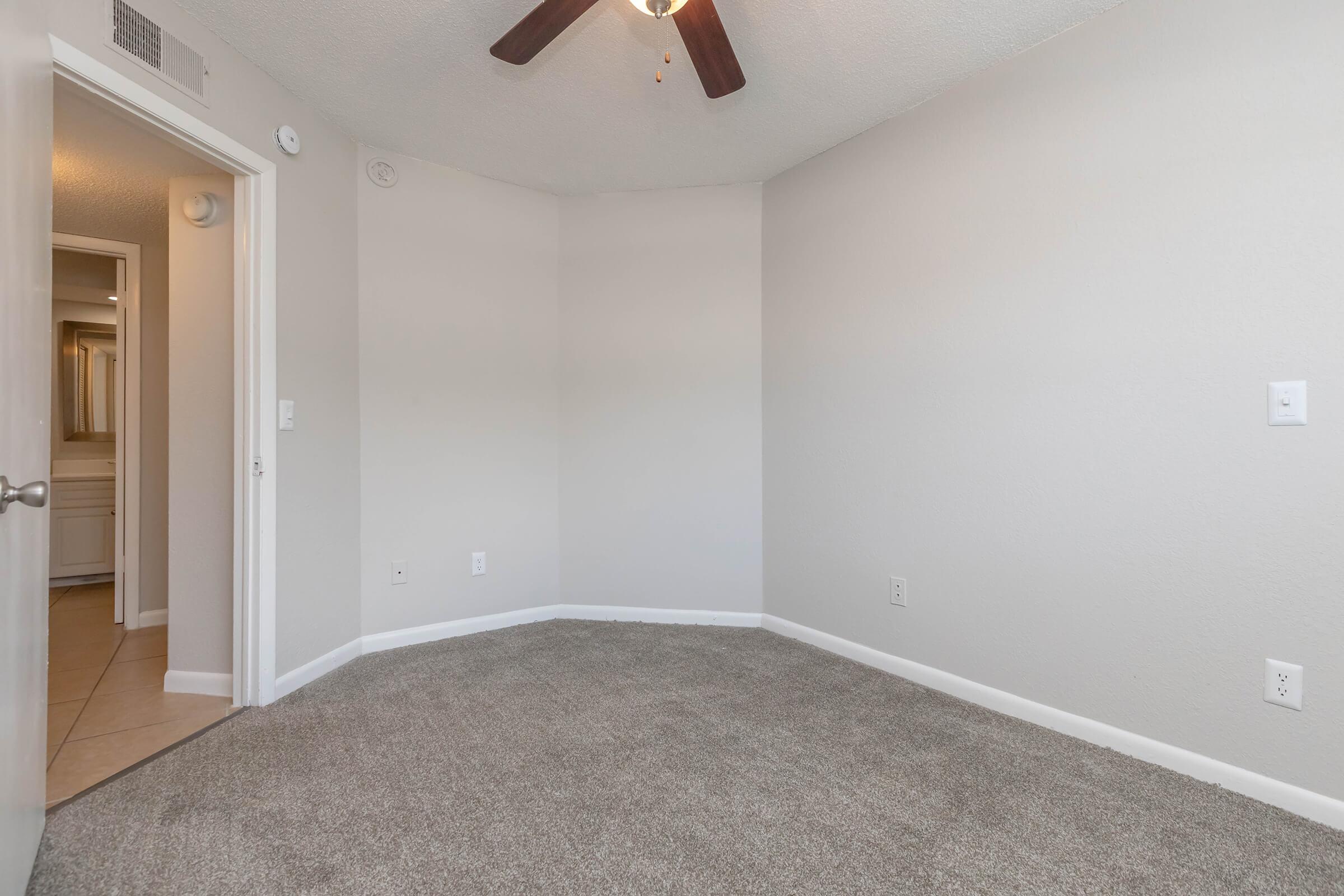 A small, empty room with beige walls and carpet, featuring a ceiling fan. There is a doorway on the left leading to a bathroom with light tiles visible. The room has no furniture and is well-lit, creating an open, airy atmosphere.