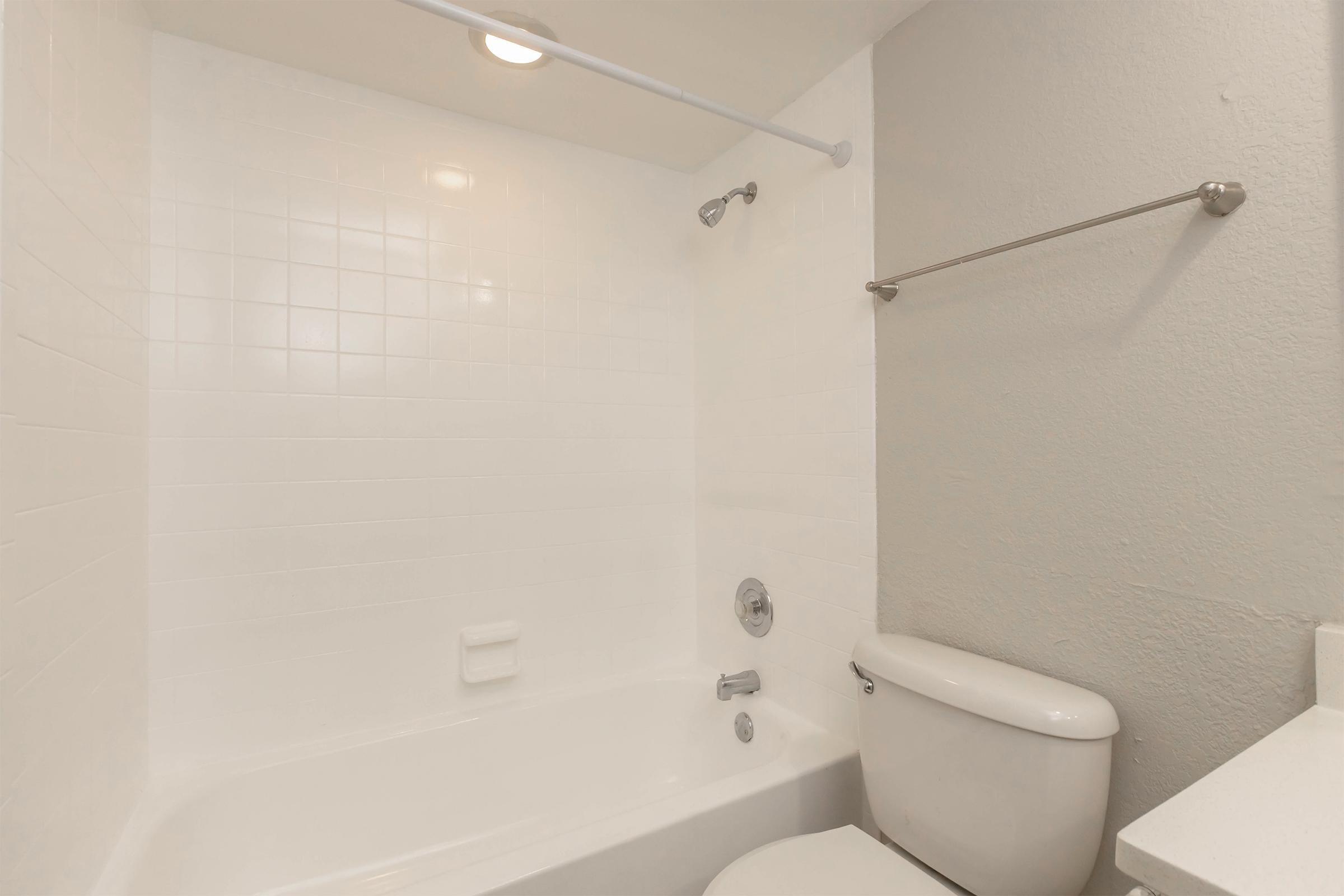 A clean and modern bathroom featuring a white tiled shower and bathtub combination, a toilet, and a simple, light-colored wall. The space is well-lit with a ceiling light and has a towel rack mounted on the wall.