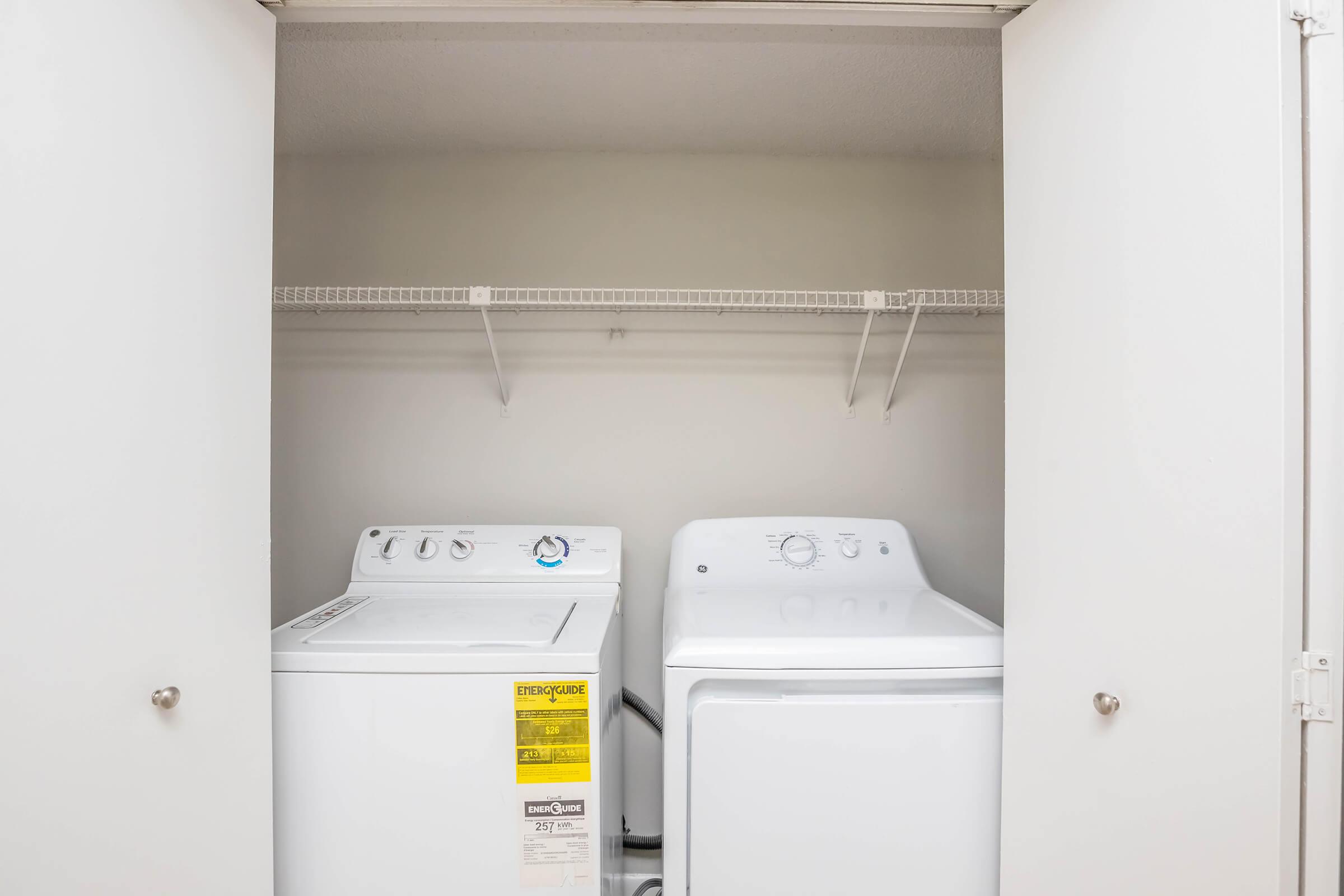 A laundry closet featuring a white top-loading washing machine and a white dryer side by side. The space is well-lit, with a simple shelving unit above for storage. The doors to the closet are open, revealing the appliances against a neutral wall.