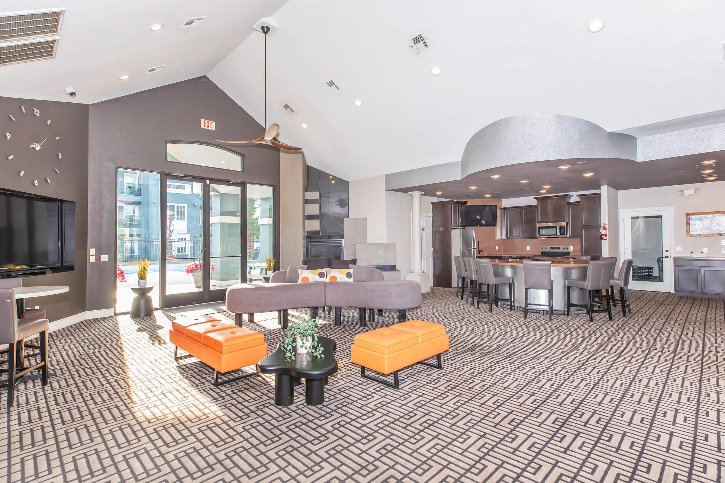 A modern, spacious lobby area featuring stylish furniture, including gray sofas, orange ottomans, and a large coffee table. There are large windows allowing natural light, and a kitchenette area with bar seating. The decor includes sleek lines and contemporary design elements, creating a welcoming atmosphere.