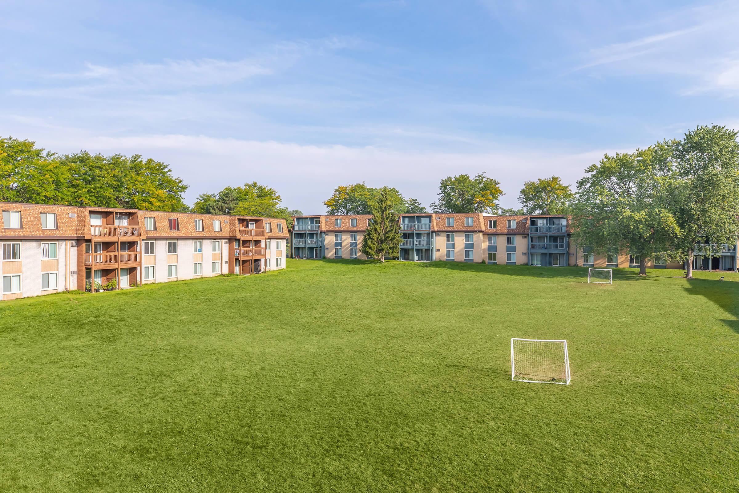 A spacious green area surrounded by residential buildings, featuring two small soccer goals on the grassy field. Trees line the perimeter, providing shade. The scene is under a clear blue sky, creating a peaceful outdoor setting for recreational activities.