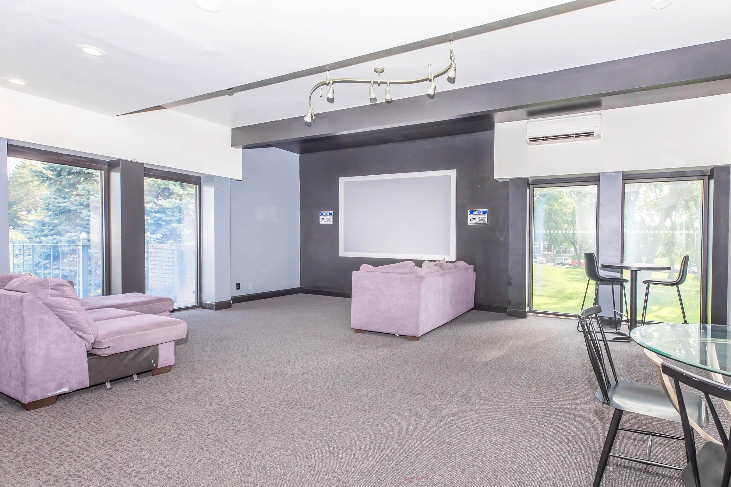 A spacious lounge area featuring two light purple sofas, a glass dining table with black chairs, and large windows allowing natural light. The walls are painted dark gray, and there is a projection screen on one wall. An air conditioning unit is visible, and the outdoor view includes greenery.