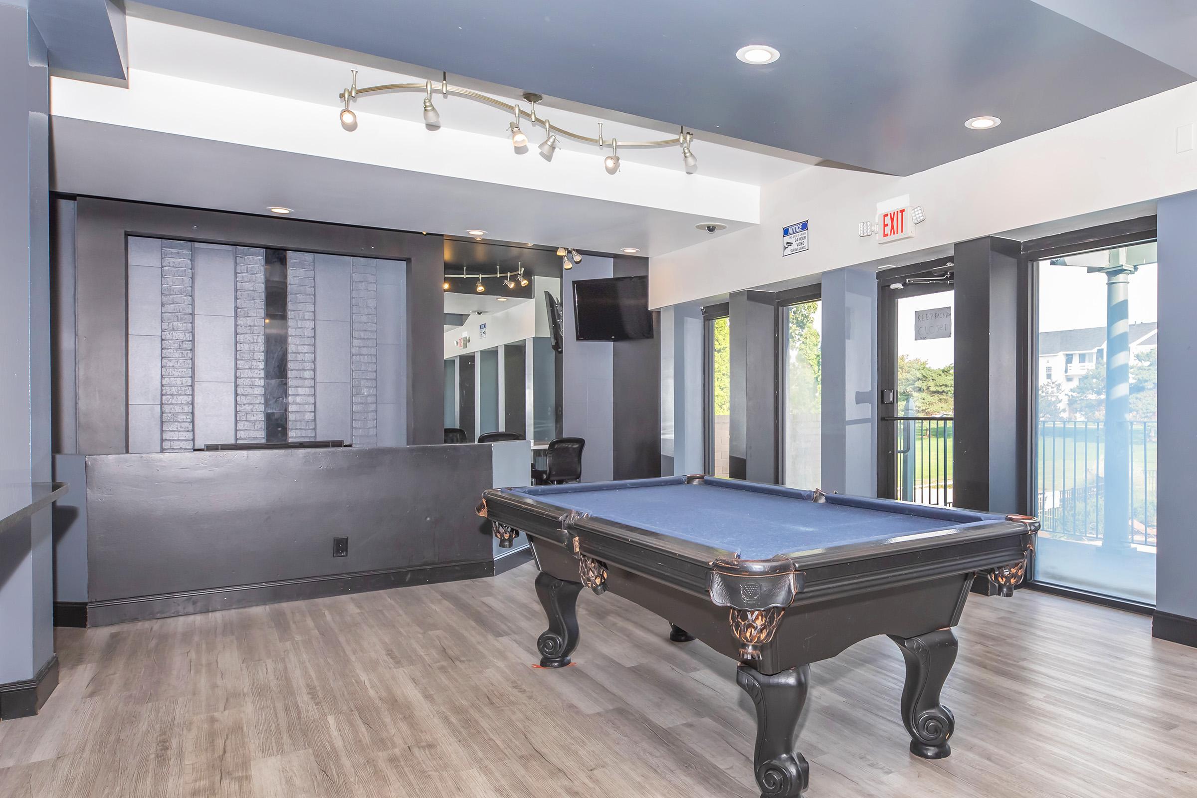 A modern indoor recreation area featuring a pool table with blue felt, dark wood accents, and a sleek design. The walls are accented with dark colors, and there are large windows providing natural light. A small bar area is visible in the background, along with seating and a TV mounted on the wall.