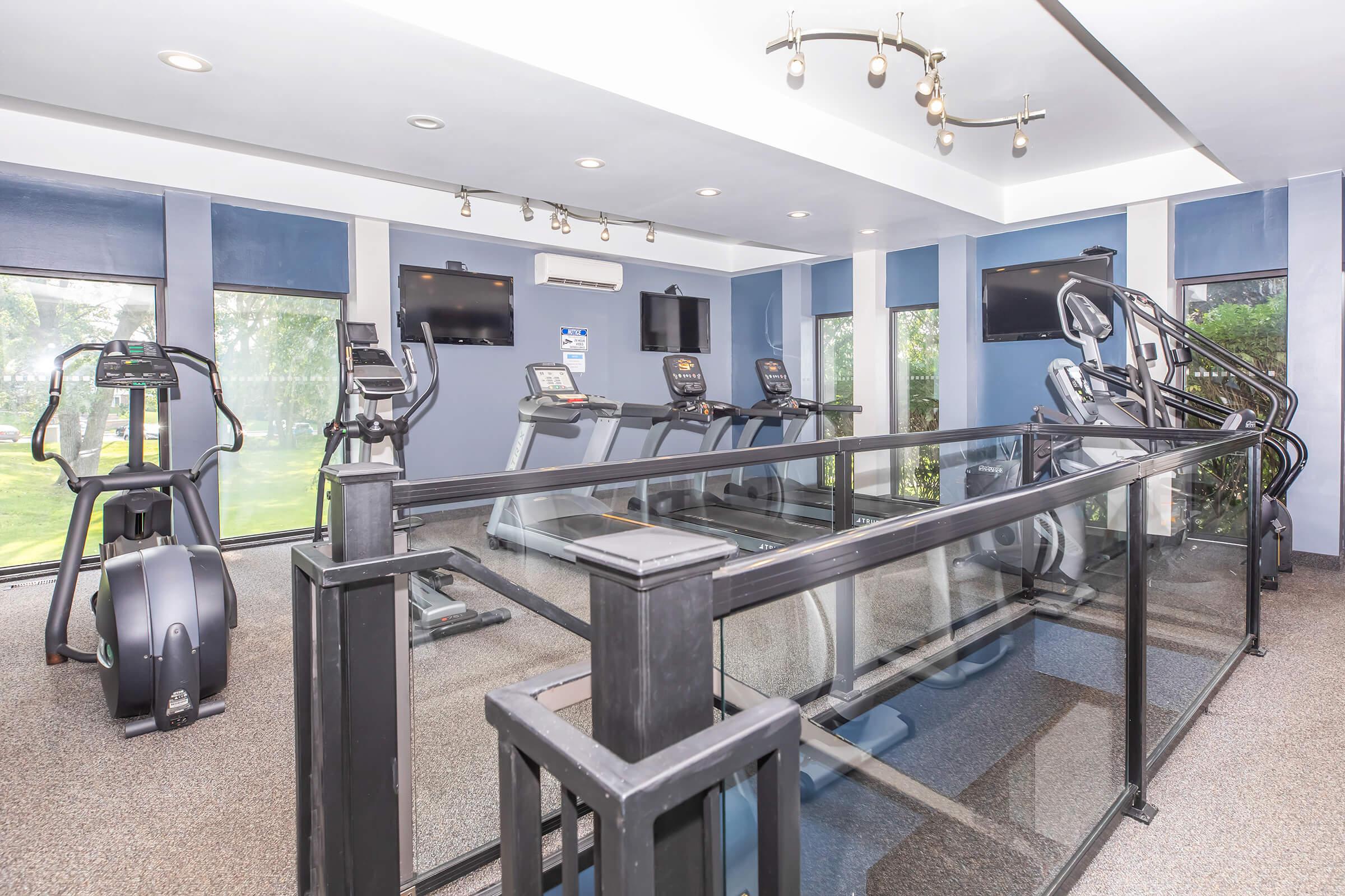 A modern fitness room featuring exercise equipment such as elliptical machines and treadmills. The space is well-lit with large windows offering a view of greenery outside. Multiple screens are visible on the walls, enhancing the workout environment. The flooring is carpeted for comfort.
