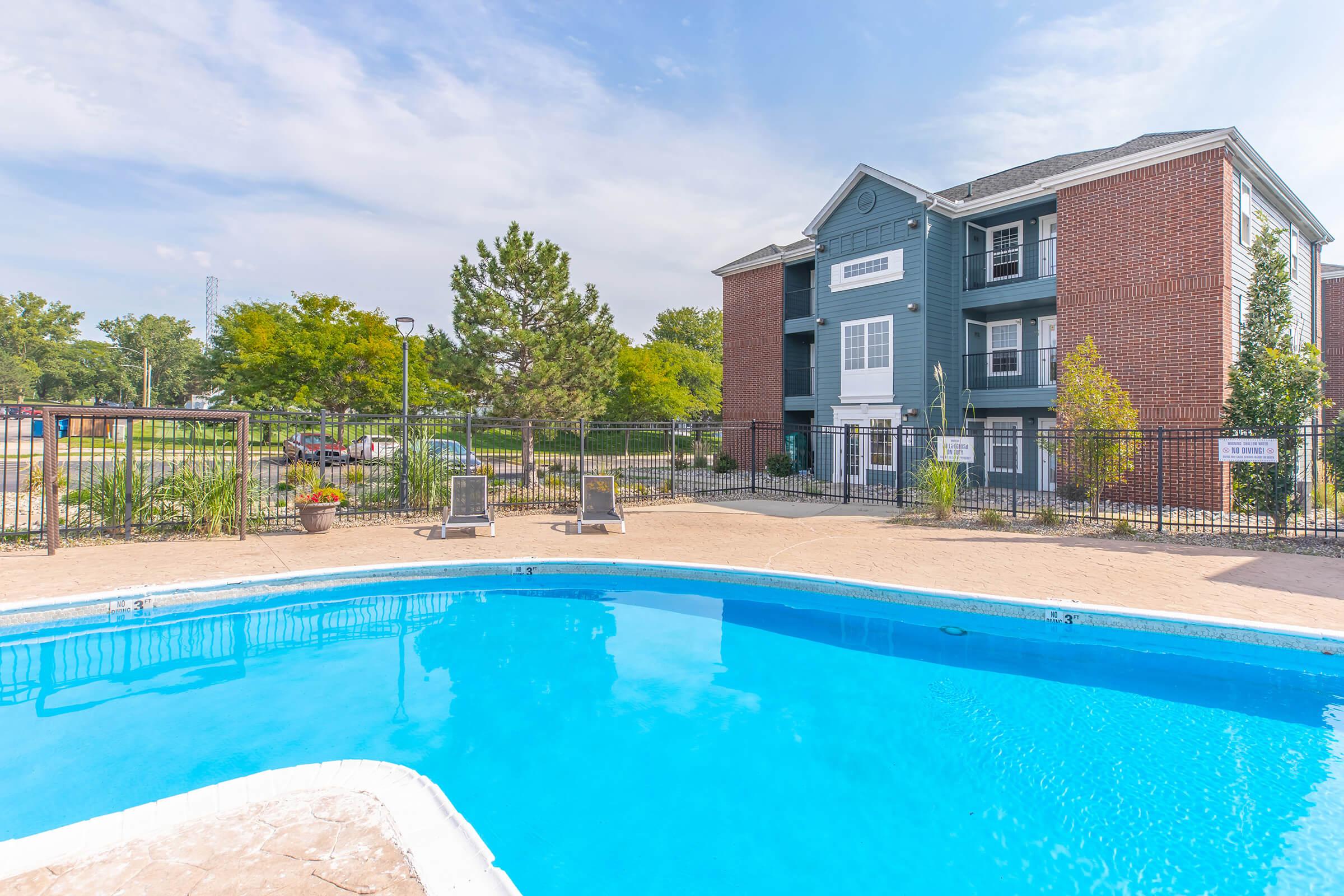 A bright blue swimming pool surrounded by a tan deck, with two lounge chairs nearby. In the background, there is a multi-story apartment building with a mix of blue and brick exterior. Lush green trees and landscaping are visible around the pool area under a clear sky.