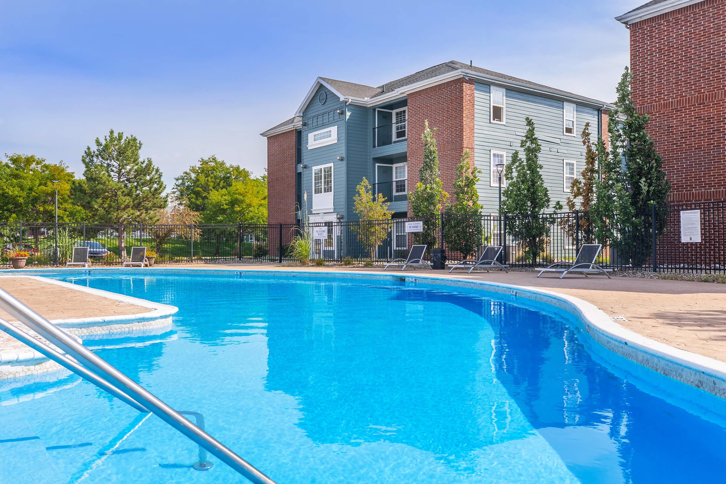A sparkling blue swimming pool with a curved shape, surrounded by lounge chairs. In the background, a multi-story residential building with a mix of brick and blue siding is visible, along with trees and greenery. The clear sky adds to the inviting atmosphere of the outdoor space.