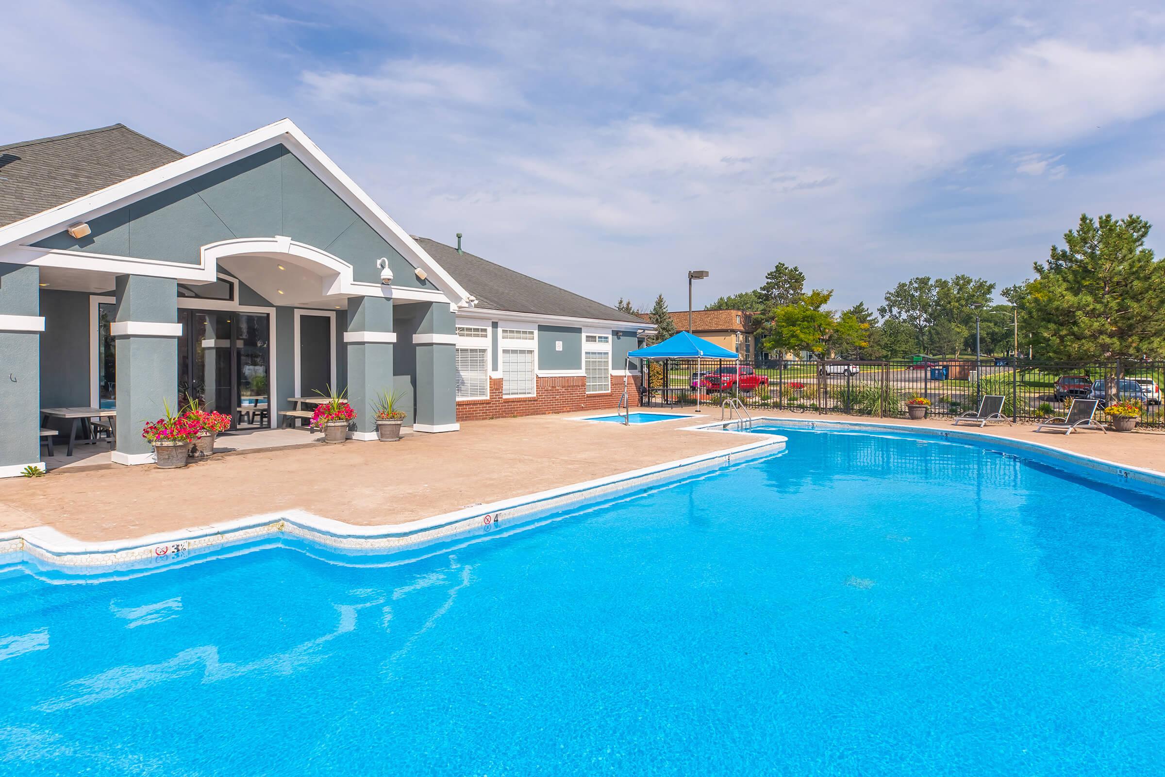 A clear blue swimming pool next to a building with large windows and a shaded outdoor seating area. Colorful flower pots adorn the patio, and lounge chairs are positioned around the pool. In the background, trees and a cloudy sky are visible, creating a relaxing outdoor atmosphere.