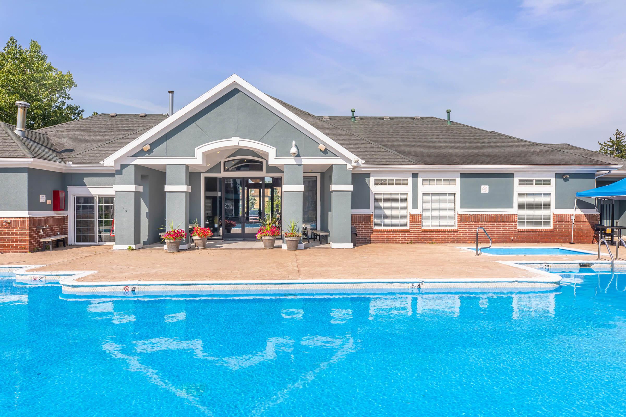 A clear blue swimming pool in front of a modern building with gray exterior and white trim. The building features large windows and an entrance flanked by colorful flower pots. A shaded area with a blue umbrella is visible beside the pool, under a blue sky with some clouds.