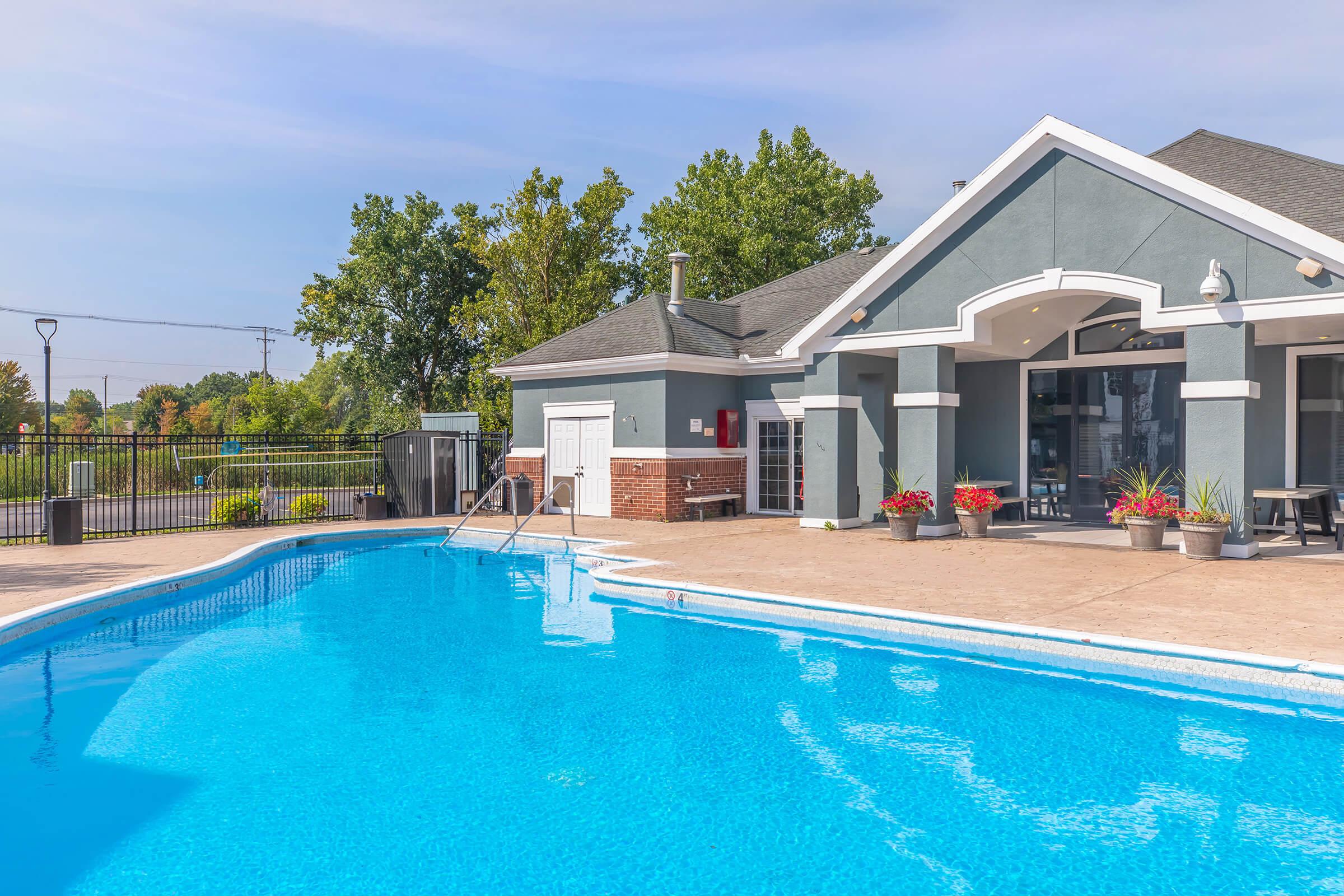 A clear blue swimming pool with a gentle curve, surrounded by a concrete deck. In the background, a modern building features large windows and a shaded entrance. Lush green trees provide natural scenery, and colorful potted plants add decoration near the pool area. The sky is bright with a few clouds.