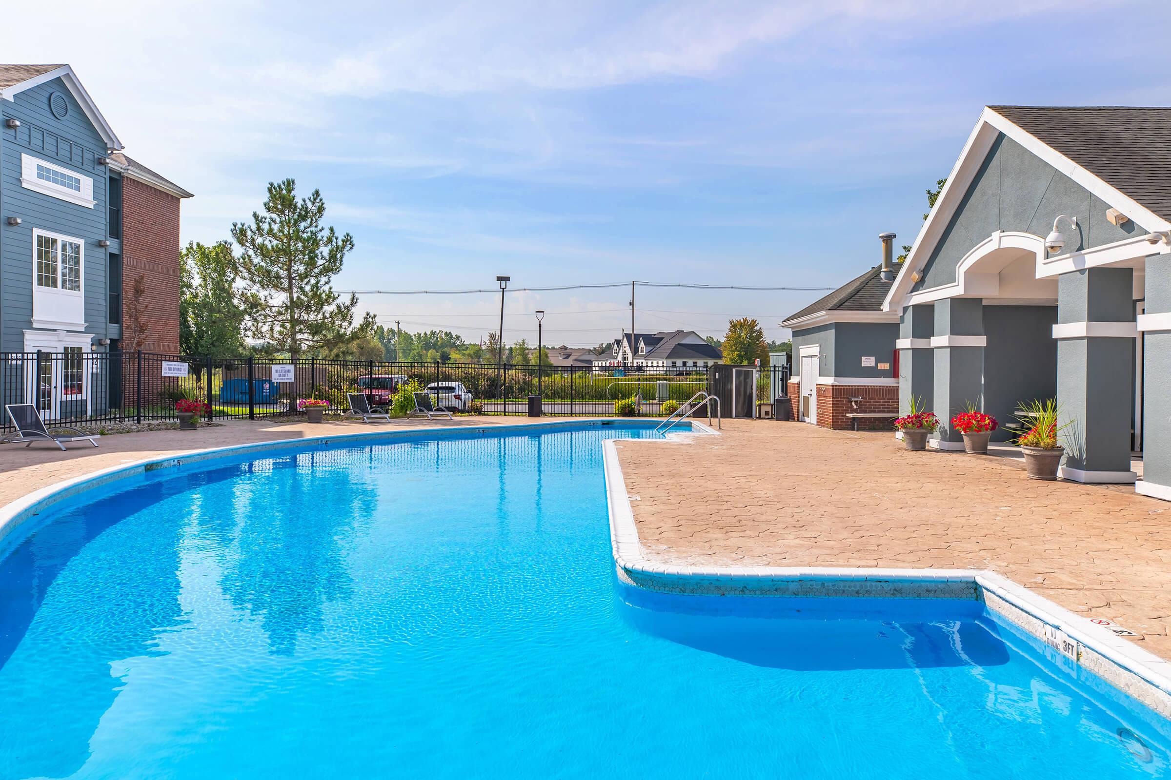 A clear, inviting swimming pool surrounded by a stone deck, with potted flowers nearby. In the background, there are two buildings, one with a blue exterior and another with a grayish design, along with trees and a fence. The sky is bright with a few clouds.