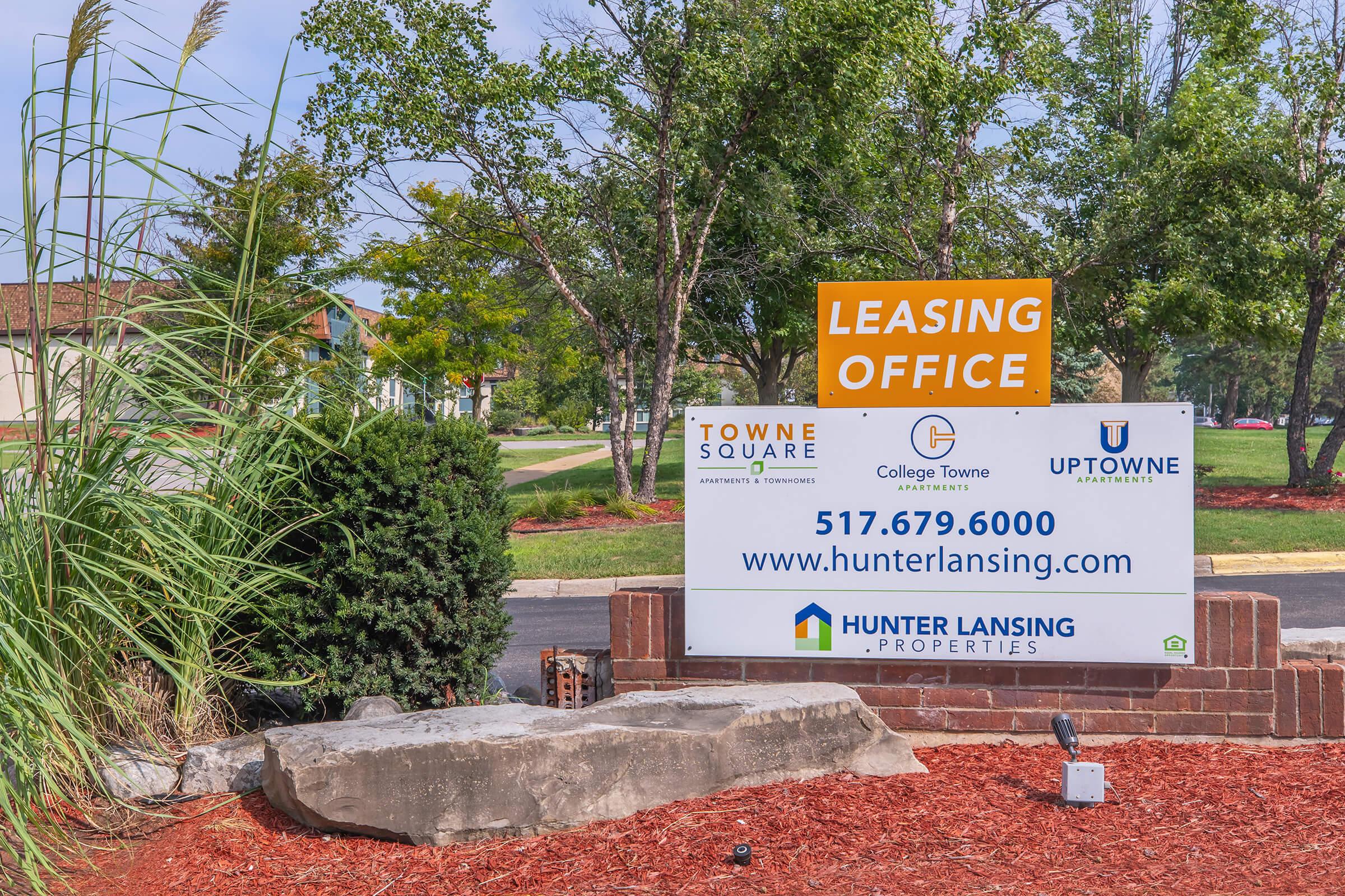 Sign for a leasing office featuring the names "Towne Square," "College Towne," and "Uptown Apartments." Includes contact information with the phone number 517.679.6000 and the company name "Hunter Lansing Properties." The sign is surrounded by landscaping elements like shrubs and stones.