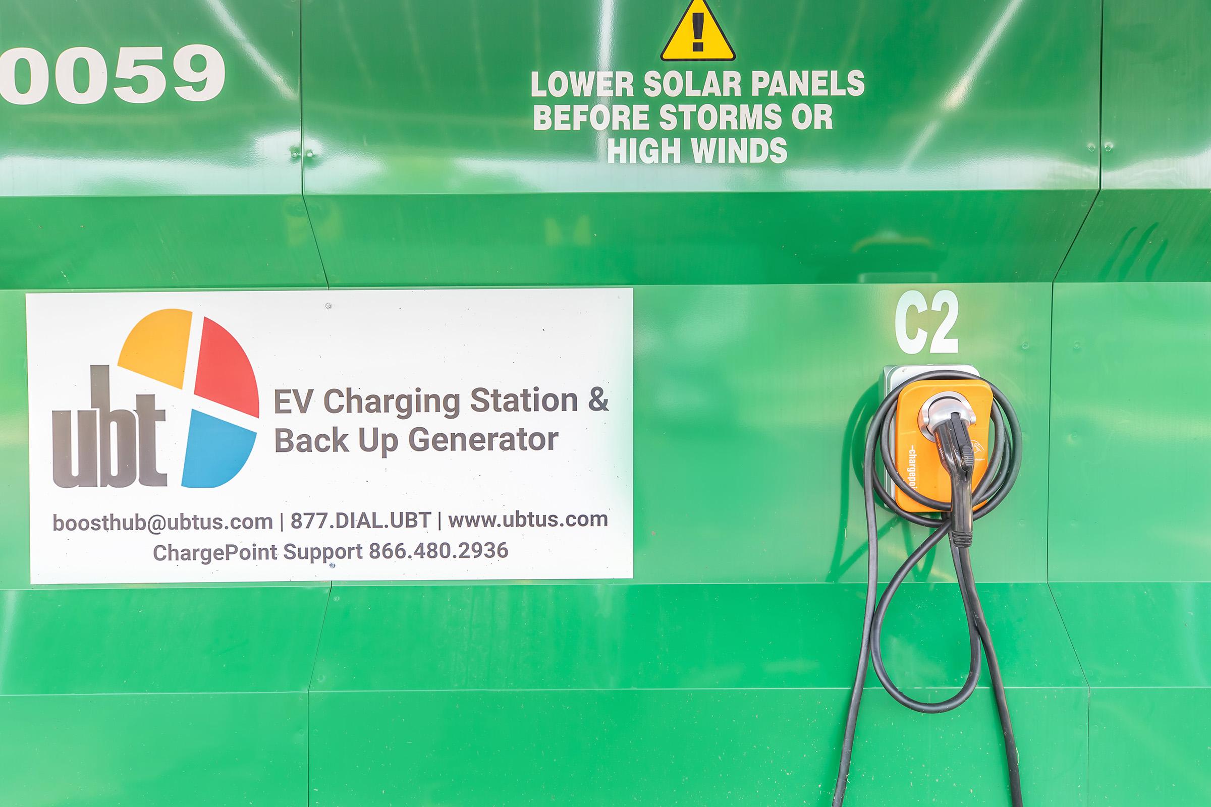 A green wall with a warning sign about lowering solar panels during storms or high winds. There is a sign for "ubt EV Charging Station & Back Up Generator" and a charging station (C2) with a charging cable attached to it. Contact information for support is also visible.