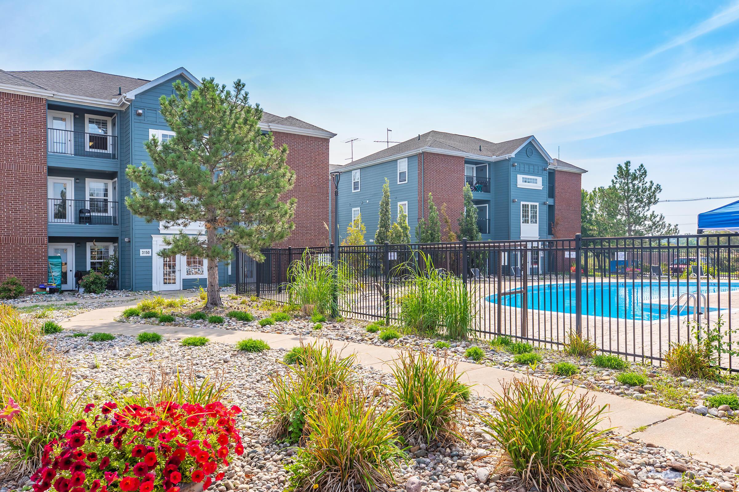 A landscaped area featuring vibrant flowers and greenery, with two multi-story apartment buildings in the background. A clear blue swimming pool is visible, surrounded by a black fence. The scene is bright and sunny, enhancing the inviting atmosphere of the residential community.