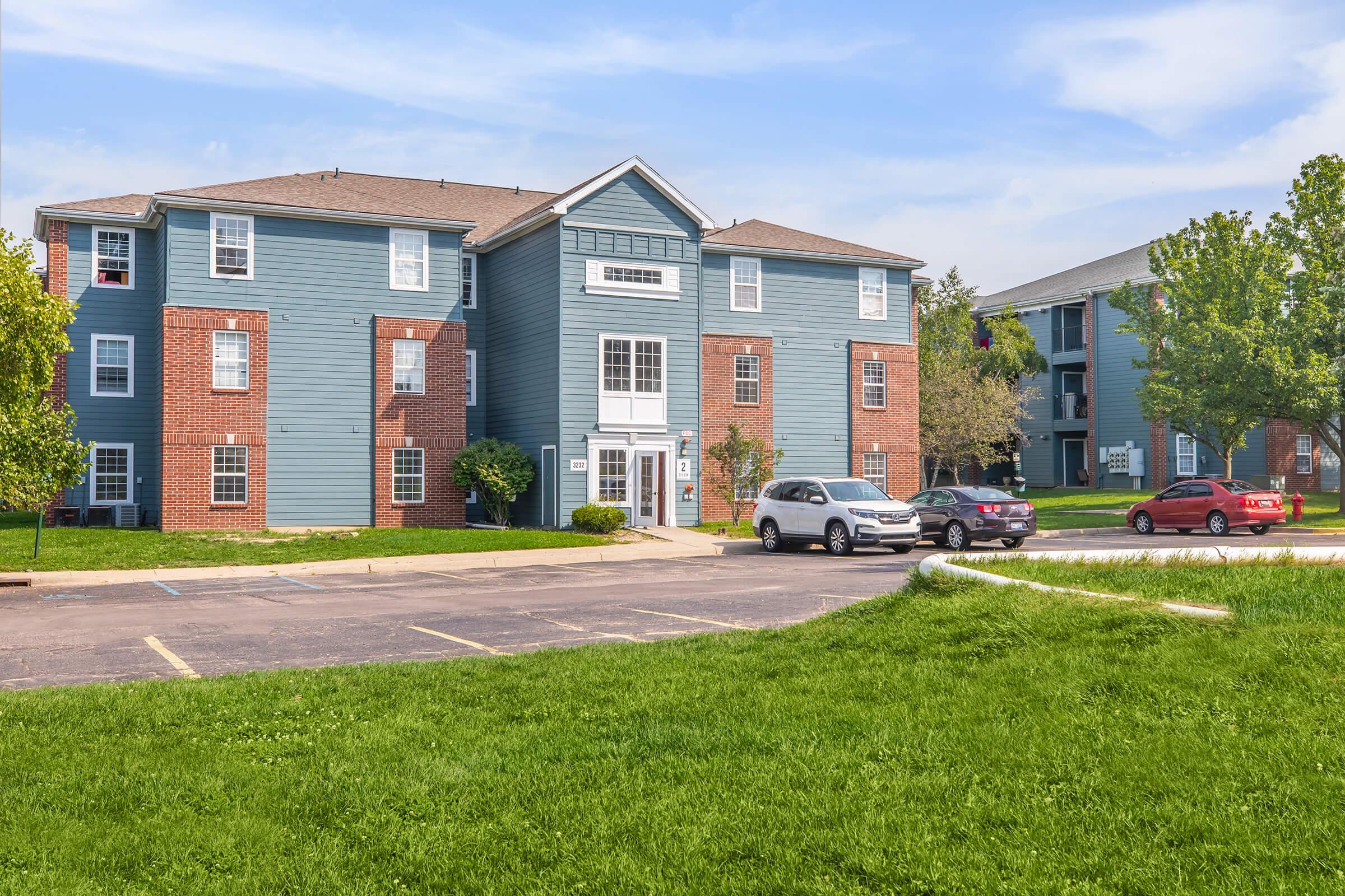 A multi-story apartment building with blue siding and red brick accents, surrounded by green grass and trees. A parking lot in the foreground has several cars parked. The sky is clear with minimal clouds, creating a bright and inviting atmosphere.