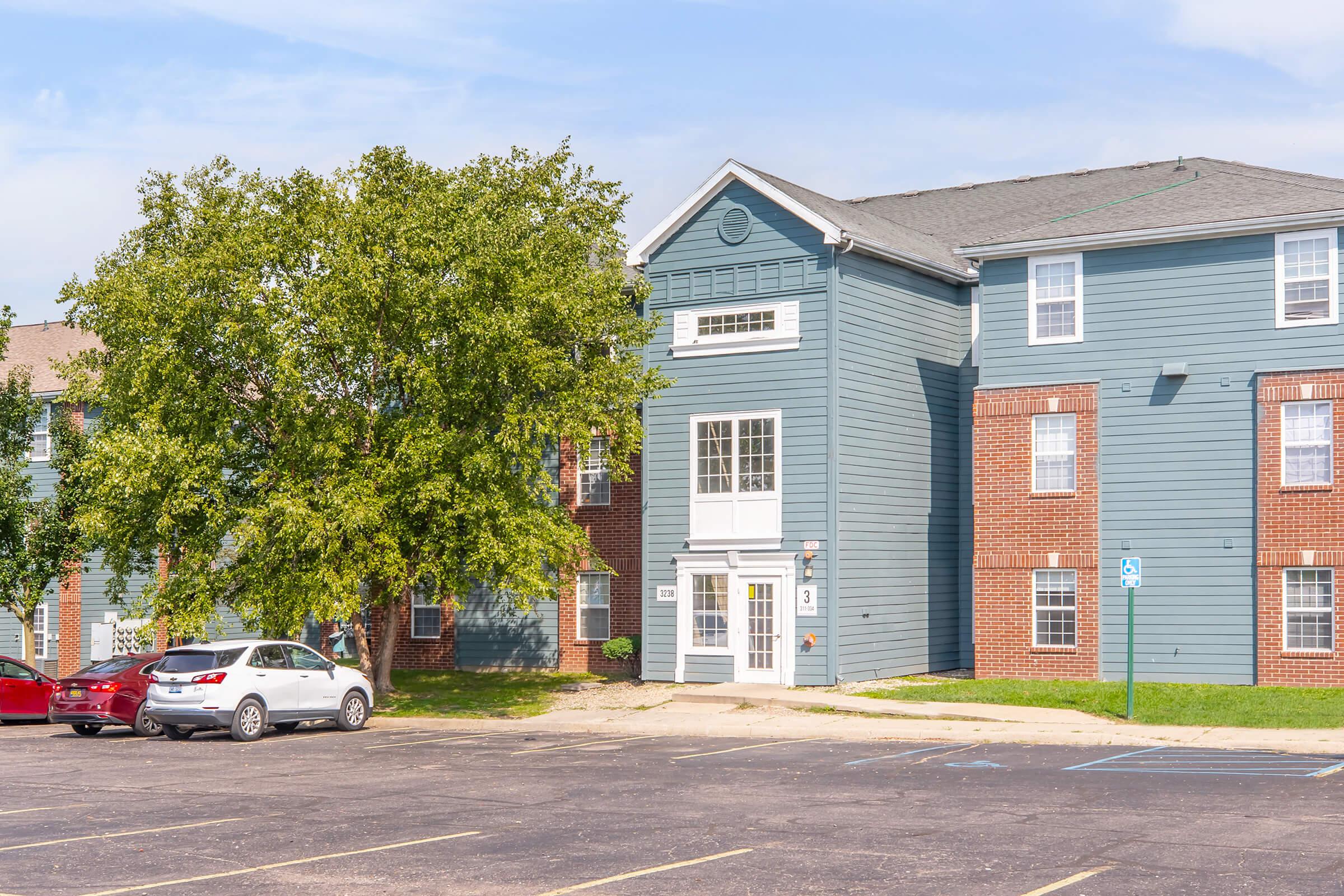 A two-story apartment building with a blue exterior and red brick accents. The entrance features a white door and large windows. Surrounding the building are parked cars and a grassy area with a tree. The parking lot shows several clear parking spaces.