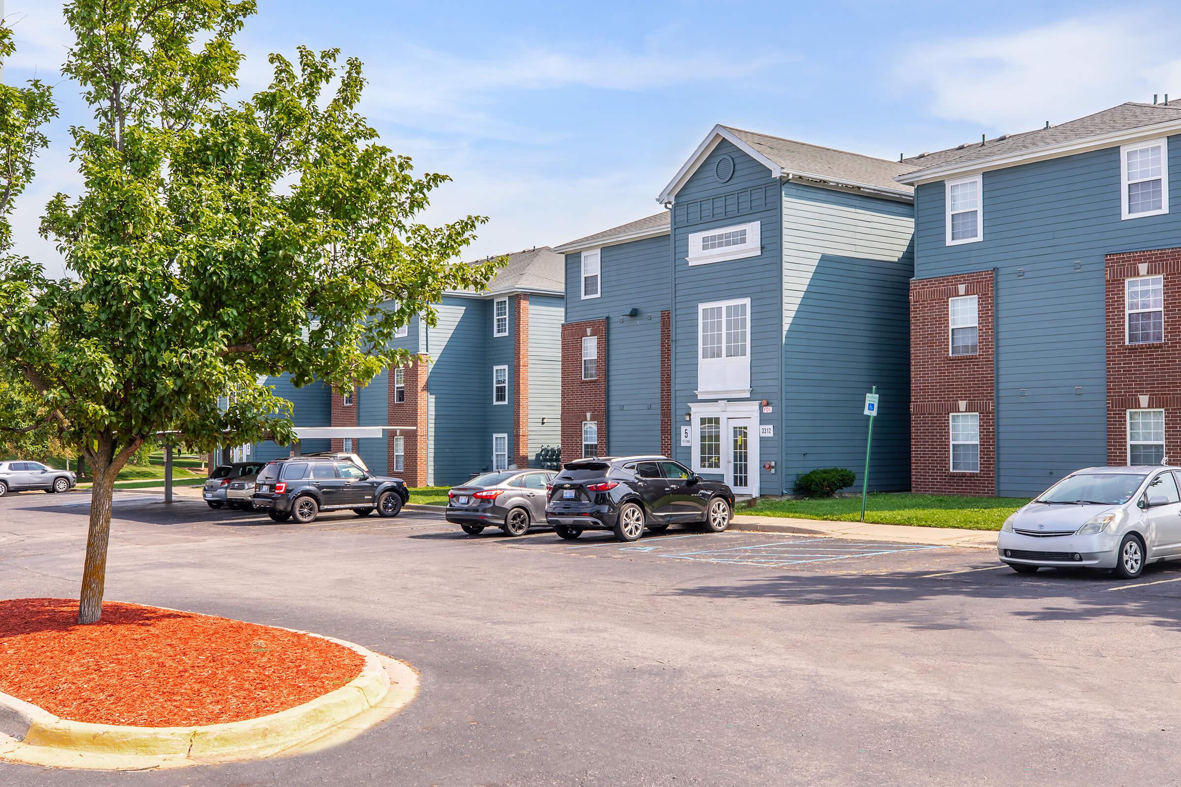 A view of a multi-story residential building with a blue exterior and brick accents. The foreground features a parking lot with several cars and a small tree. Bright red mulch is visible along the edge of the parking area, under a clear blue sky.