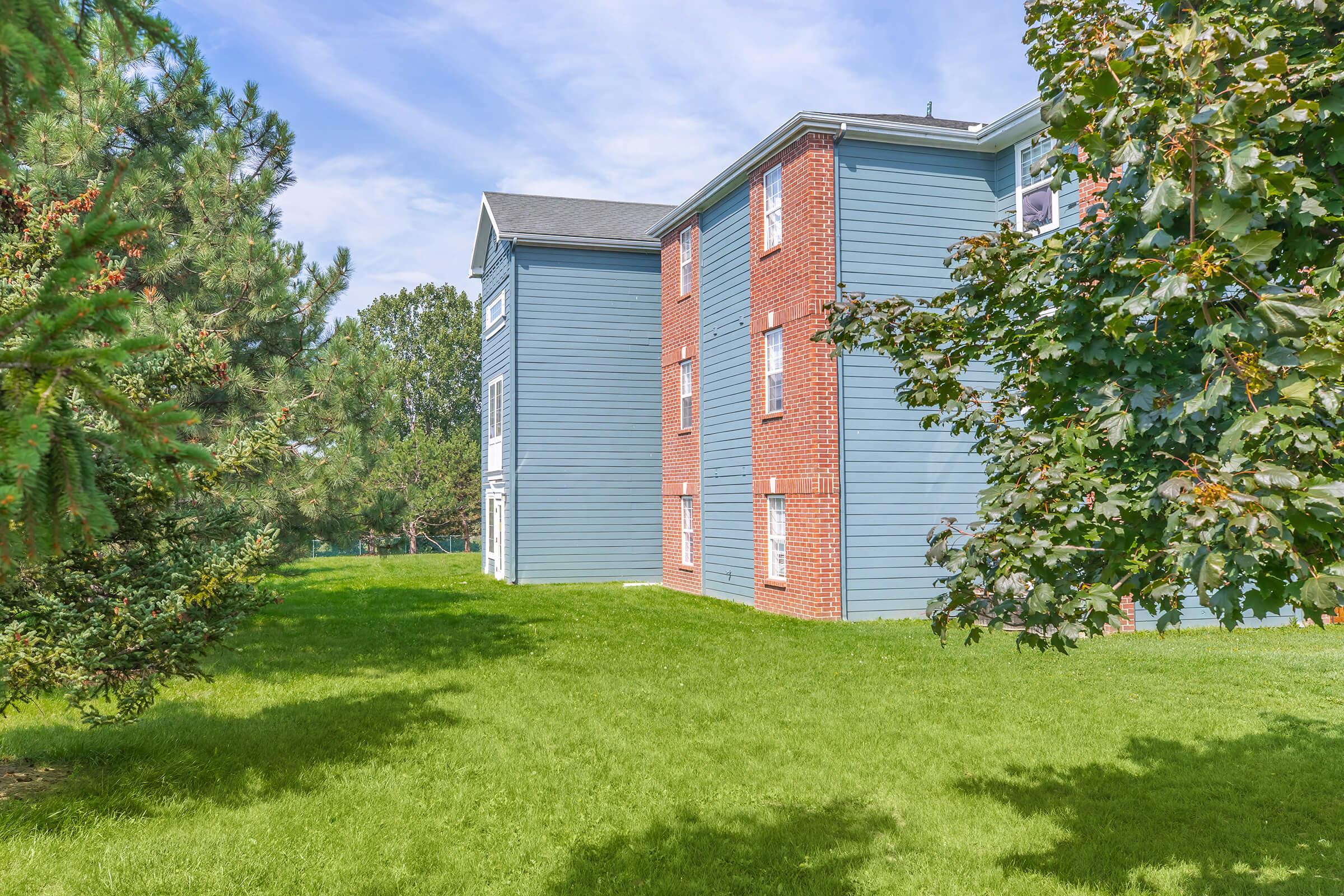 A multi-story building with a blue and red exterior, surrounded by greenery. The building has several windows and is situated on a lawn with trees nearby. The sky is partly cloudy, providing a pleasant atmosphere.
