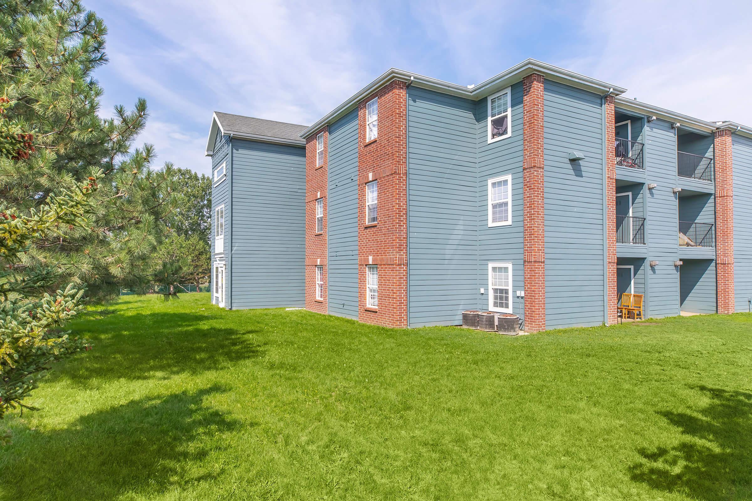 A multi-story apartment building with blue siding and red brick accents, surrounded by lush green grass and trees. The building features several windows and balconies, and there are chairs visible on the balcony of the lower level. The sky is clear with a few clouds.