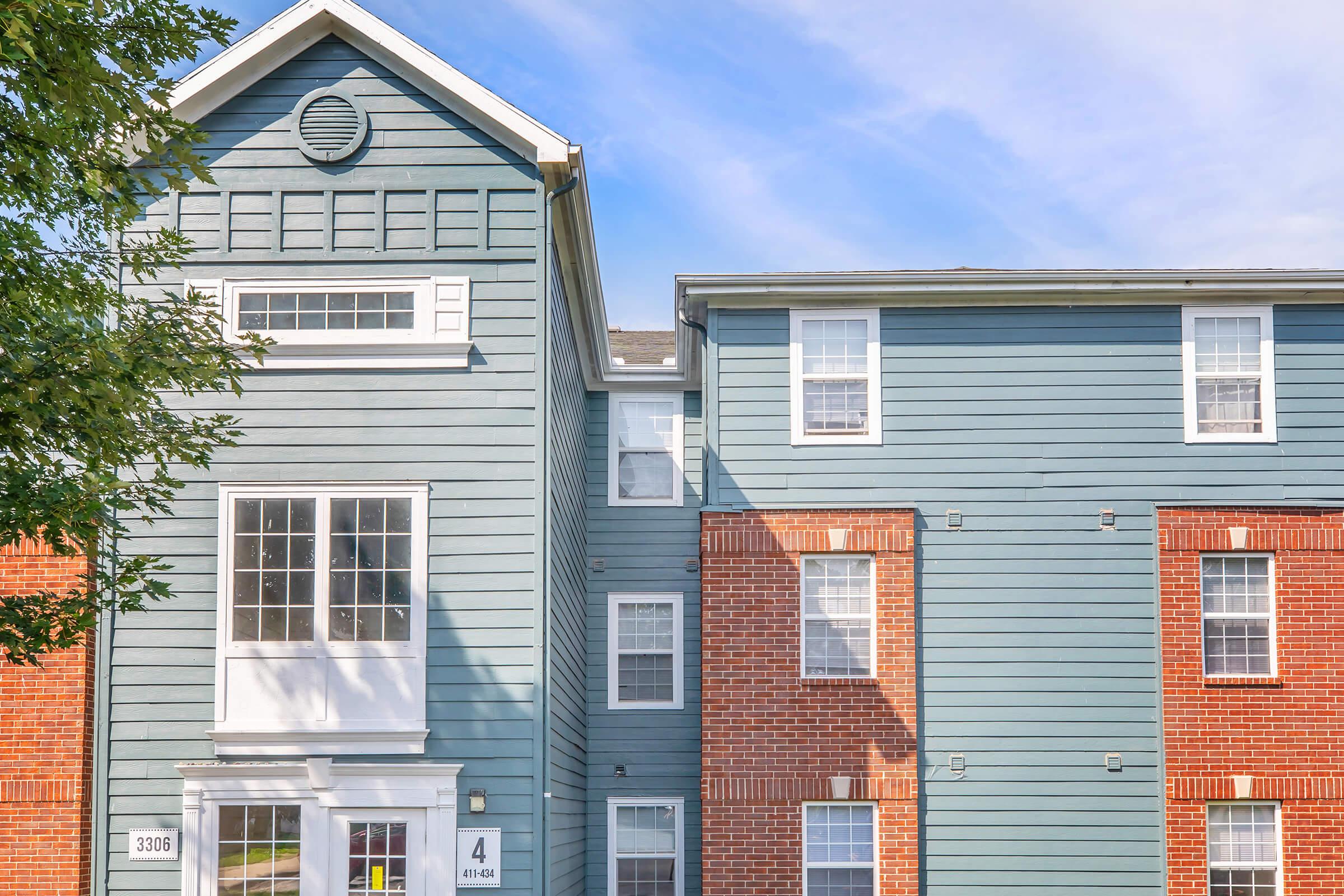 Exterior view of a multi-story residential building featuring a blue wooden facade and red brick accents. The structure includes large windows and a prominent entrance with a white door, along with numbered signs indicating apartment units. Trees partially frame the building against a clear blue sky.