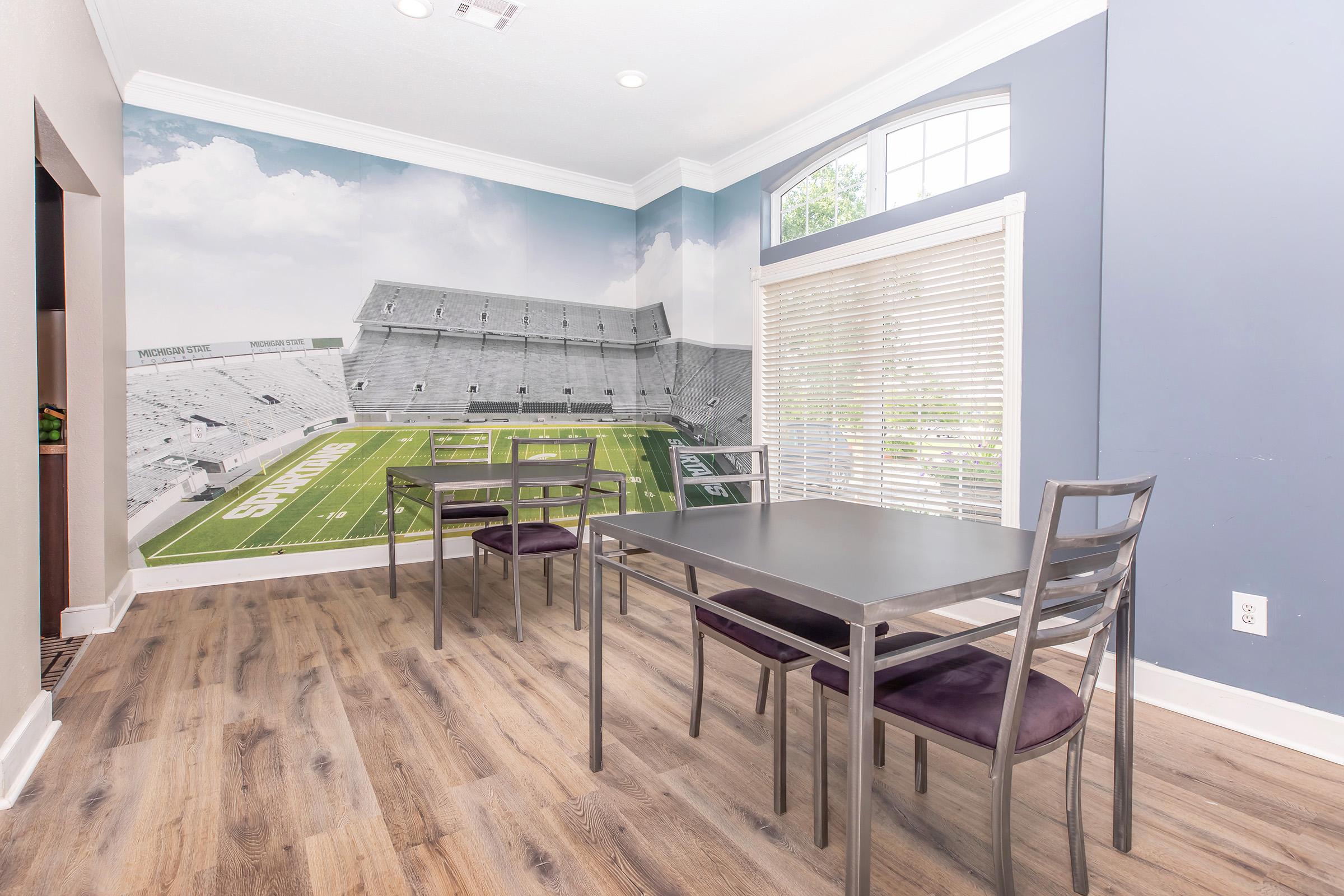 A modern dining area featuring a wall mural of a football stadium. The room has a sleek wooden floor, large windows with natural light, a rectangular dining table with four metal chairs, and a stylish design that combines sports imagery with contemporary decor.