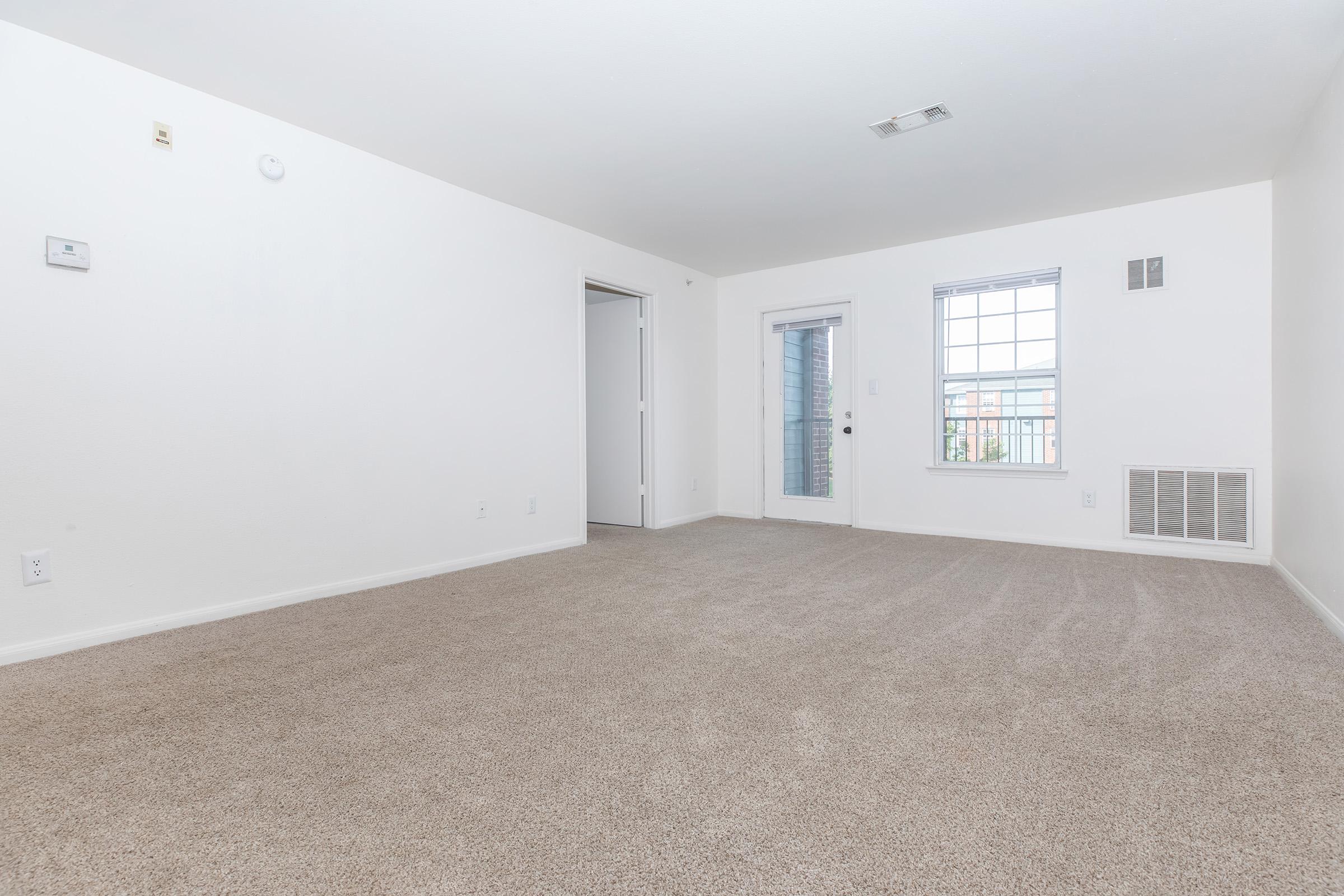 A spacious, empty living room featuring light-colored walls and carpet flooring. There is a doorway leading to another room, a sliding glass door with a window for natural light, and a wall vent. The room appears bright and clean, ready for furnishing.