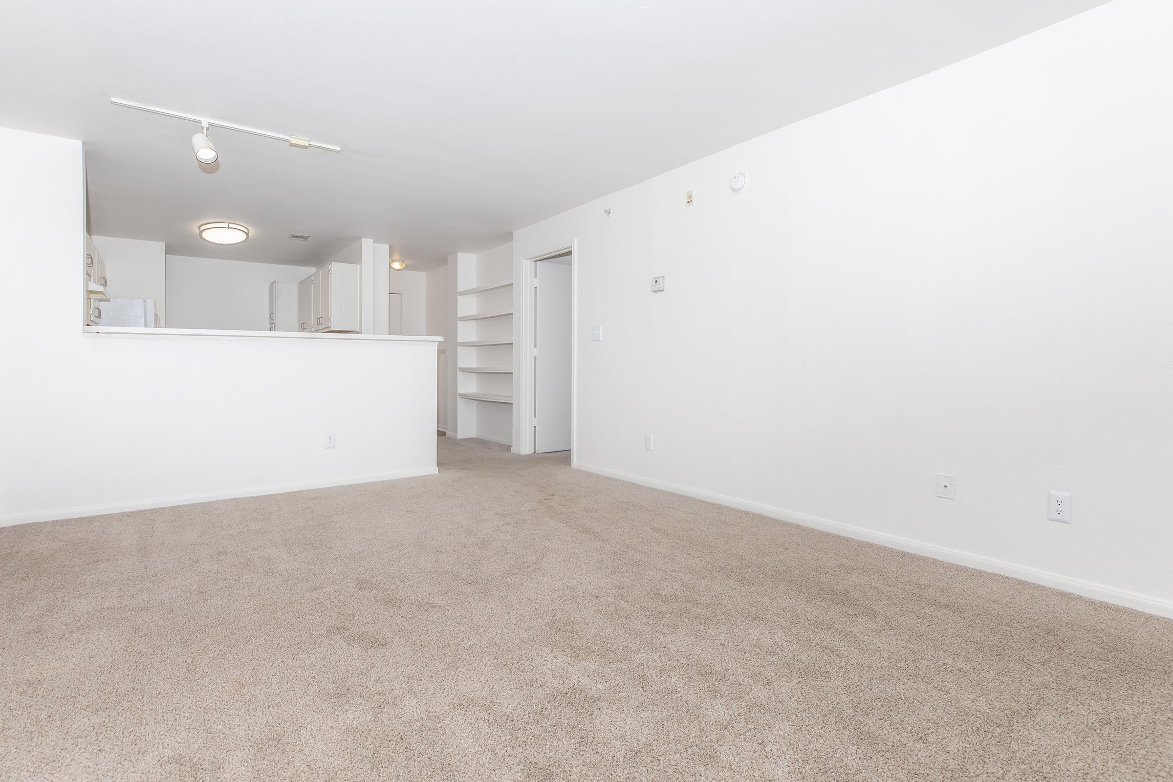 A spacious, empty room with light beige carpet and white walls. In the background, there is a partial view of a kitchen area with an open layout and a light fixture on the ceiling. A doorway leads to another room, and there are built-in shelves along one wall. The overall atmosphere is bright and inviting.