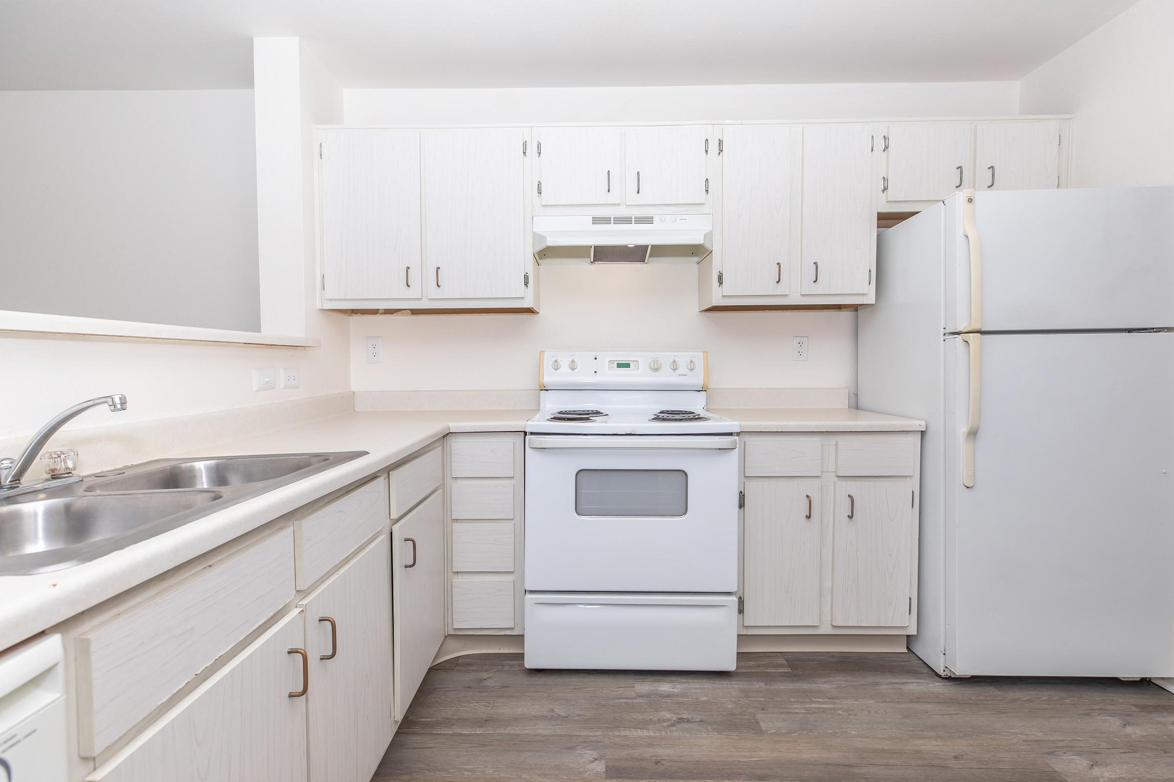 A bright and clean kitchen featuring white cabinets, a stove with an oven, a hood vent, and a refrigerator. The countertop is light-colored, and there are double sinks. The flooring is a light wood laminate, creating a modern and inviting space.