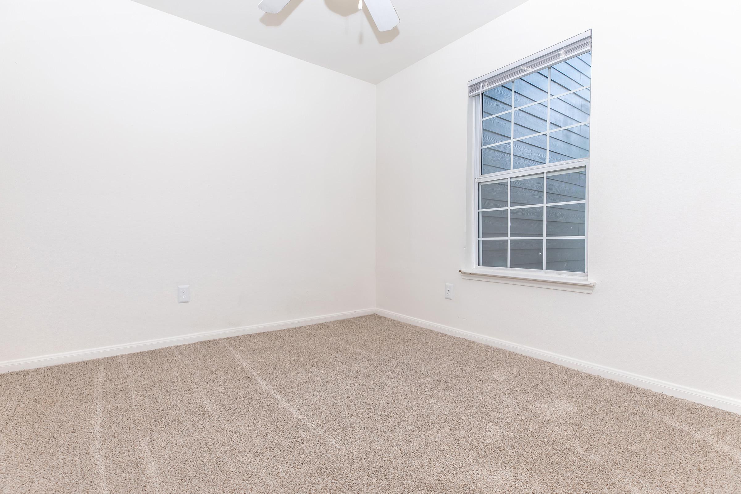A sparse, empty room with light-colored walls and beige carpet. A window with multiple panes allows natural light to enter, casting a soft glow. The ceiling features a white ceiling fan, and there are no furnishings or decorations, creating a minimalist and open atmosphere.