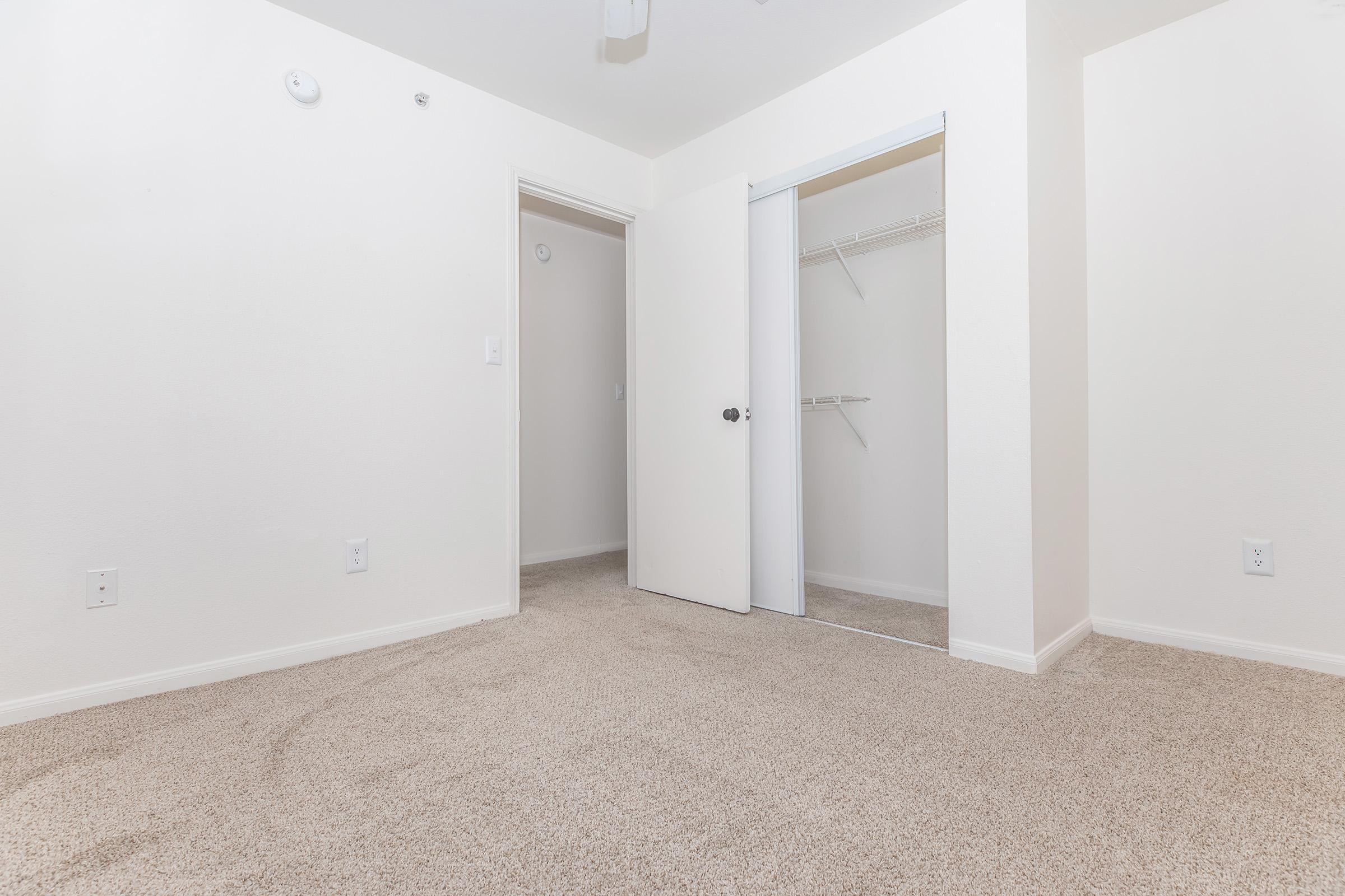 A neatly organized room featuring beige carpet, white walls, and two doors—one leading to a closet with mirrored sliding doors and the other to another room. The space is well-lit and minimalistic, providing a clean and airy atmosphere.