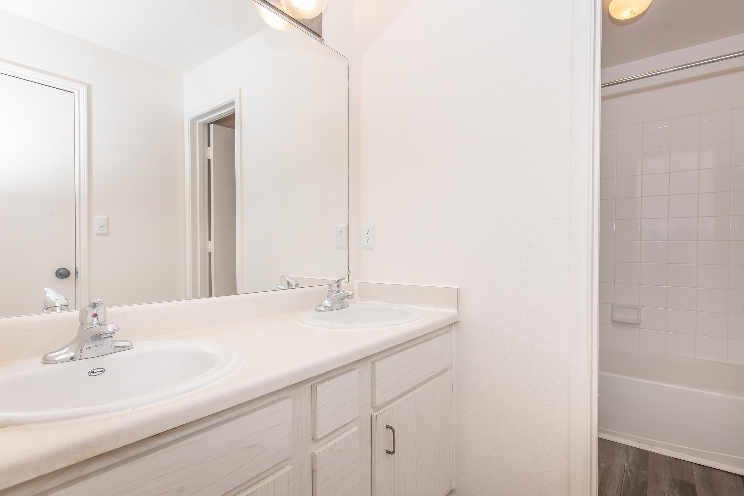 A clean, modern bathroom featuring a double sink vanity with chrome faucets, a large mirror, and light-colored walls. A shower or bathtub area is visible in the background, along with a doorway leading to another room. Natural light illuminates the space, creating a bright atmosphere.