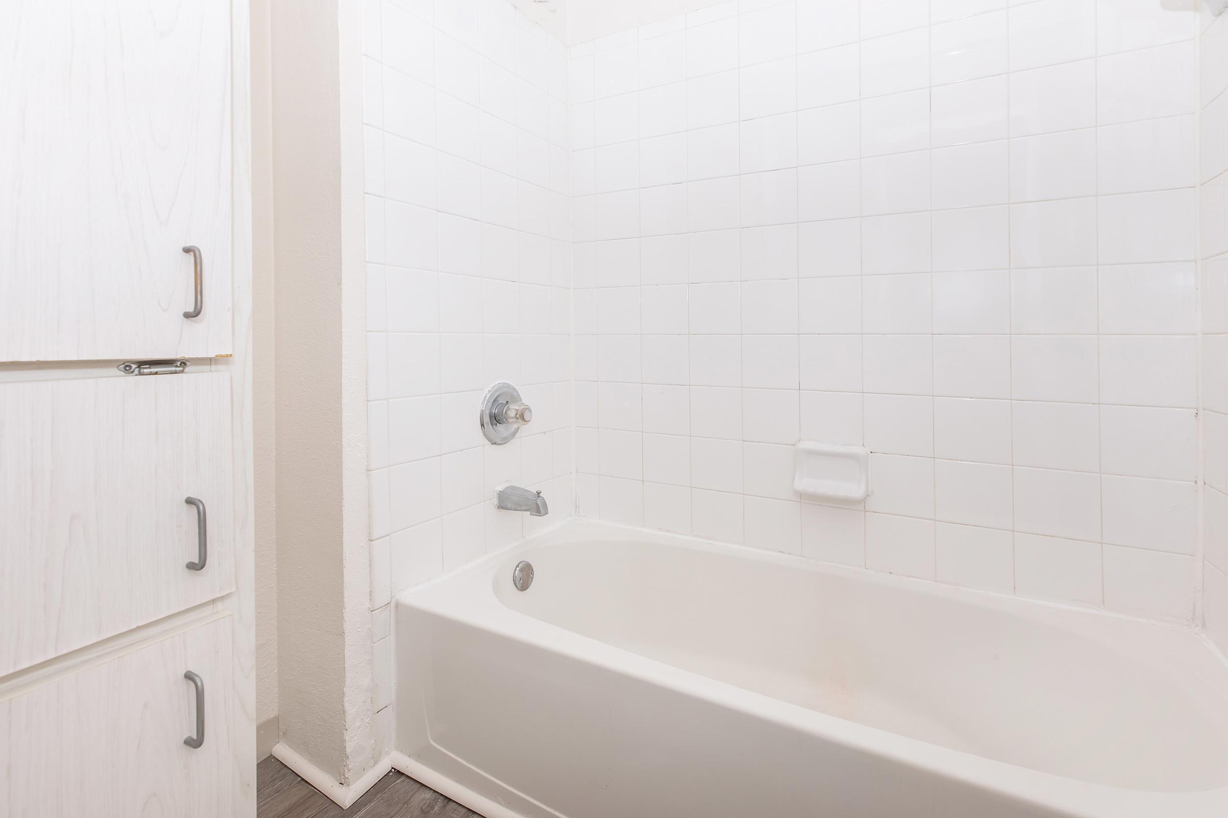 A clean, white tiled bathroom featuring a bathtub with a chrome faucet and a shower head. The wall next to the bathtub is tiled, while the floor appears to be a darker material. There is a light-colored cabinet with drawers on the left side of the image. The overall atmosphere is bright and minimalist.