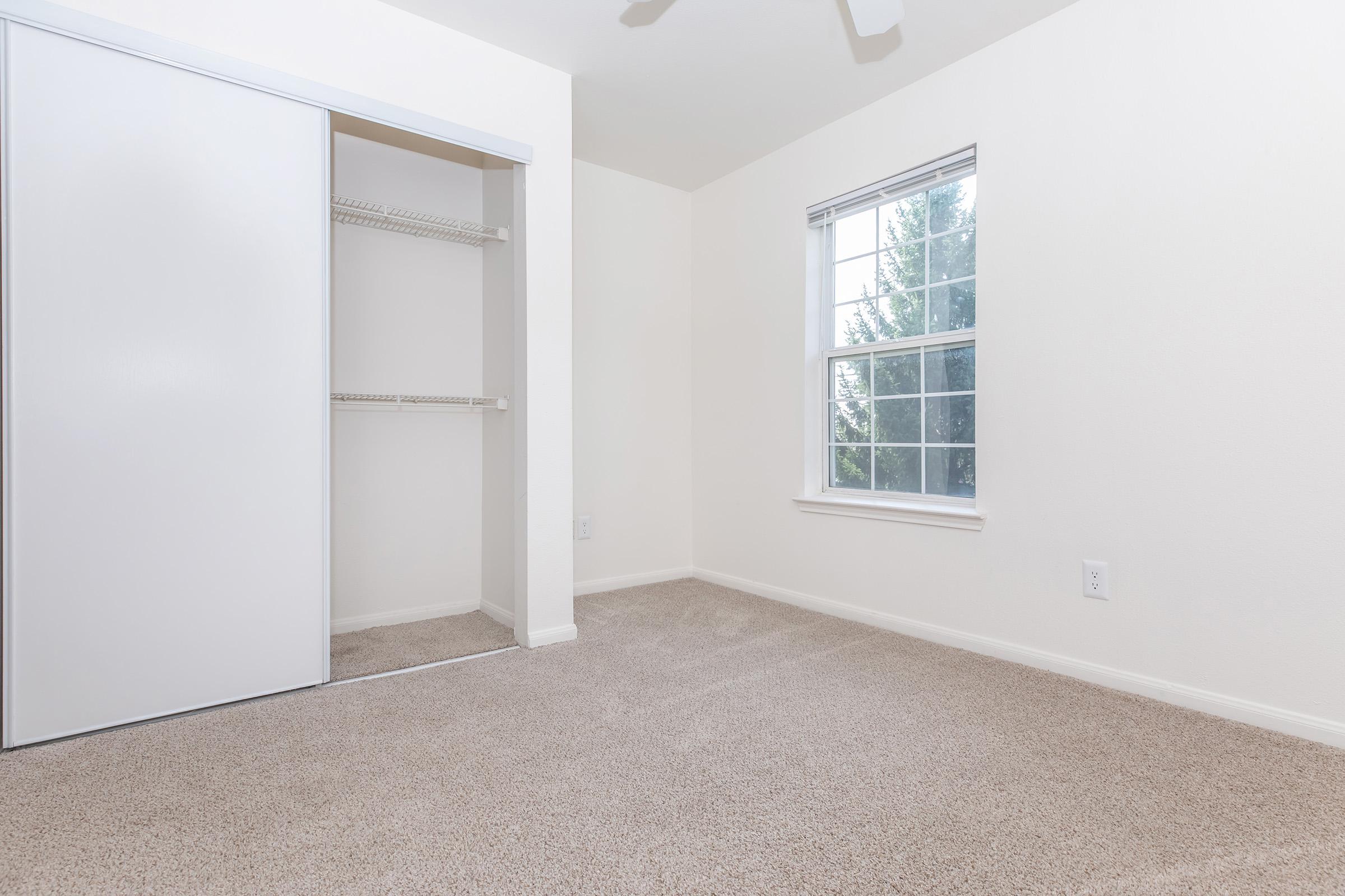 Empty room with beige carpet, white walls, and a window letting in natural light. A closed closet with sliding doors is on the left, and the room is unadorned, creating a spacious feel.