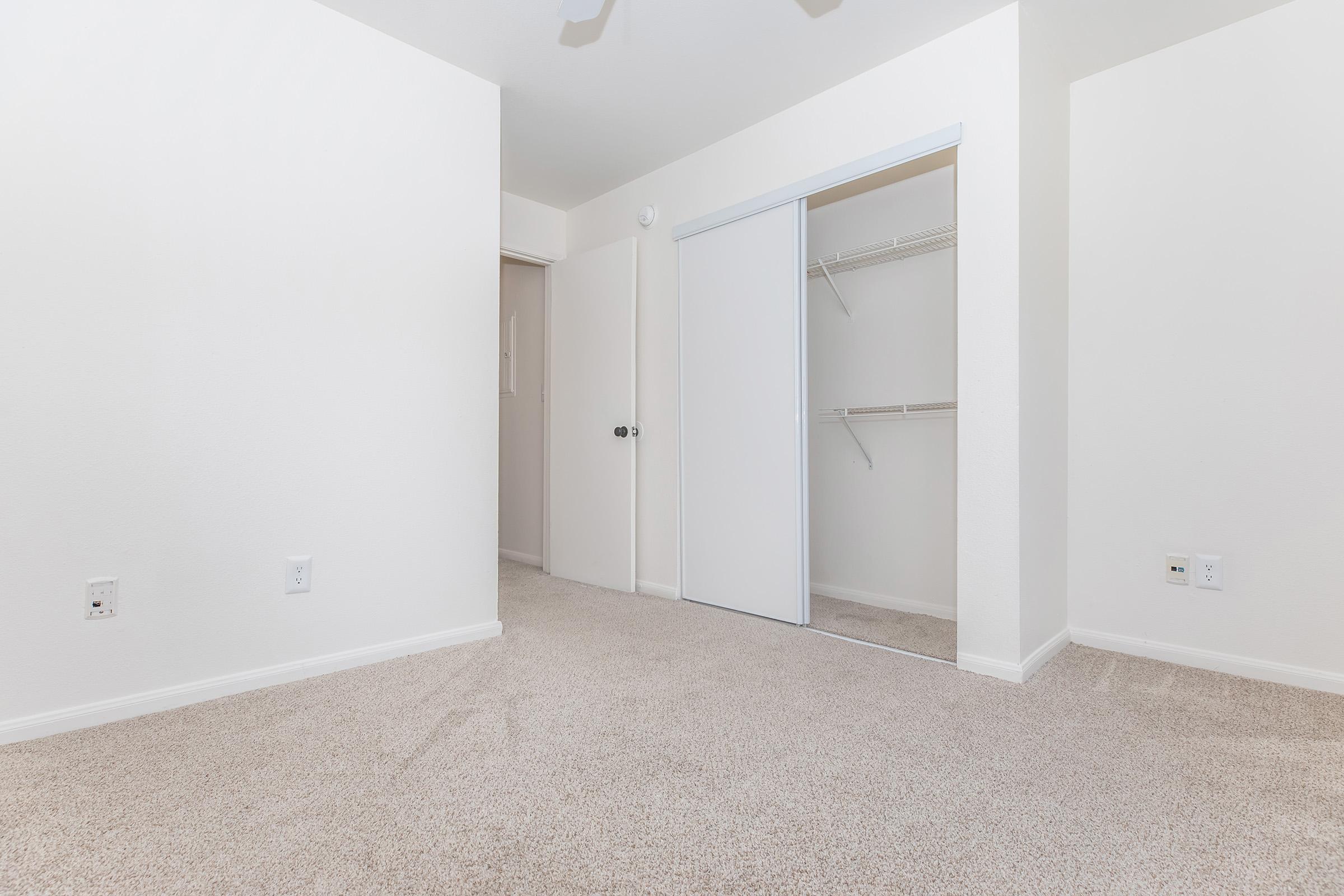 A spacious, empty room with light-colored walls and carpeted flooring. There’s a closet with sliding doors on one side and a doorway leading to another area. The room has a clean, minimalistic look, making it suitable for various uses or decorations.