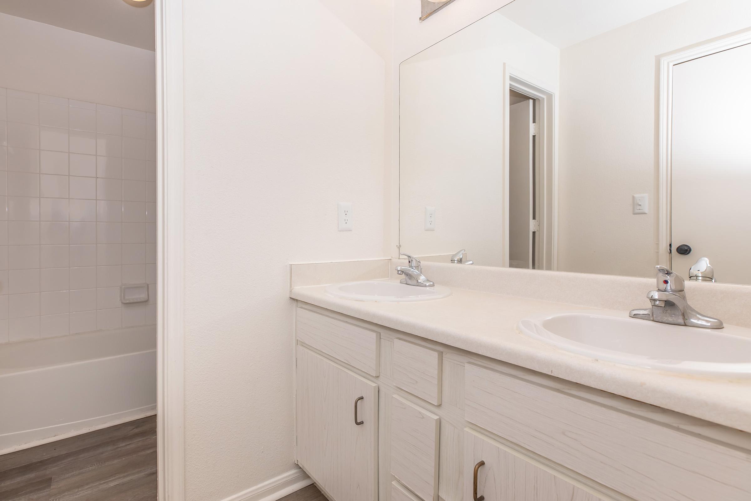 A clean bathroom featuring a double vanity with two sinks, mirrors above, and light-colored countertops. Next to the vanity is a bathtub with a tiled wall, and a door leading to another room. The walls are painted in a neutral tone, creating a bright and spacious feel.