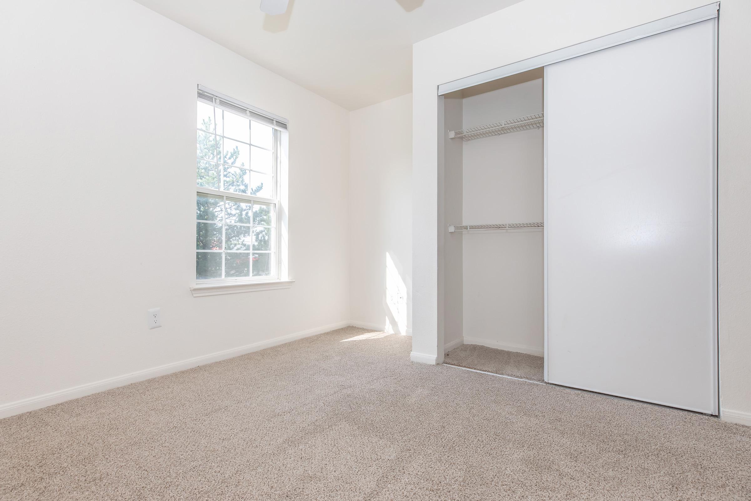 A well-lit, empty bedroom featuring beige carpet, a single window with natural light, and a closet with sliding doors. The walls are painted a light color, and the overall space appears clean and ready for decoration.