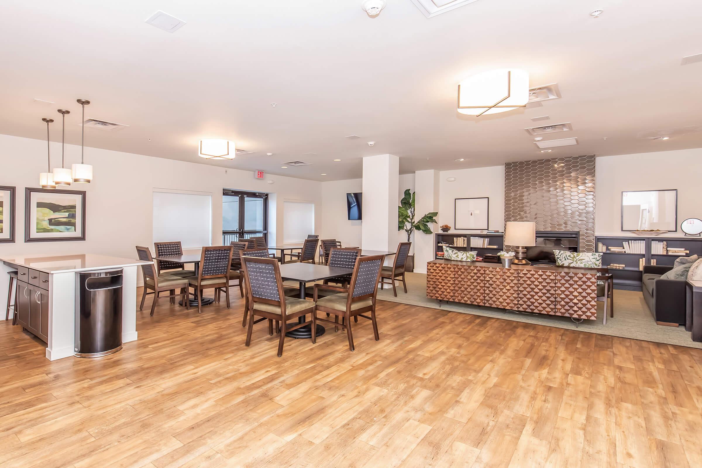 A spacious, modern common area featuring wooden flooring, multiple tables with chairs, and a cozy seating arrangement. The decor includes contemporary light fixtures, artwork on the walls, and plants, creating an inviting atmosphere for residents or guests to relax and socialize.