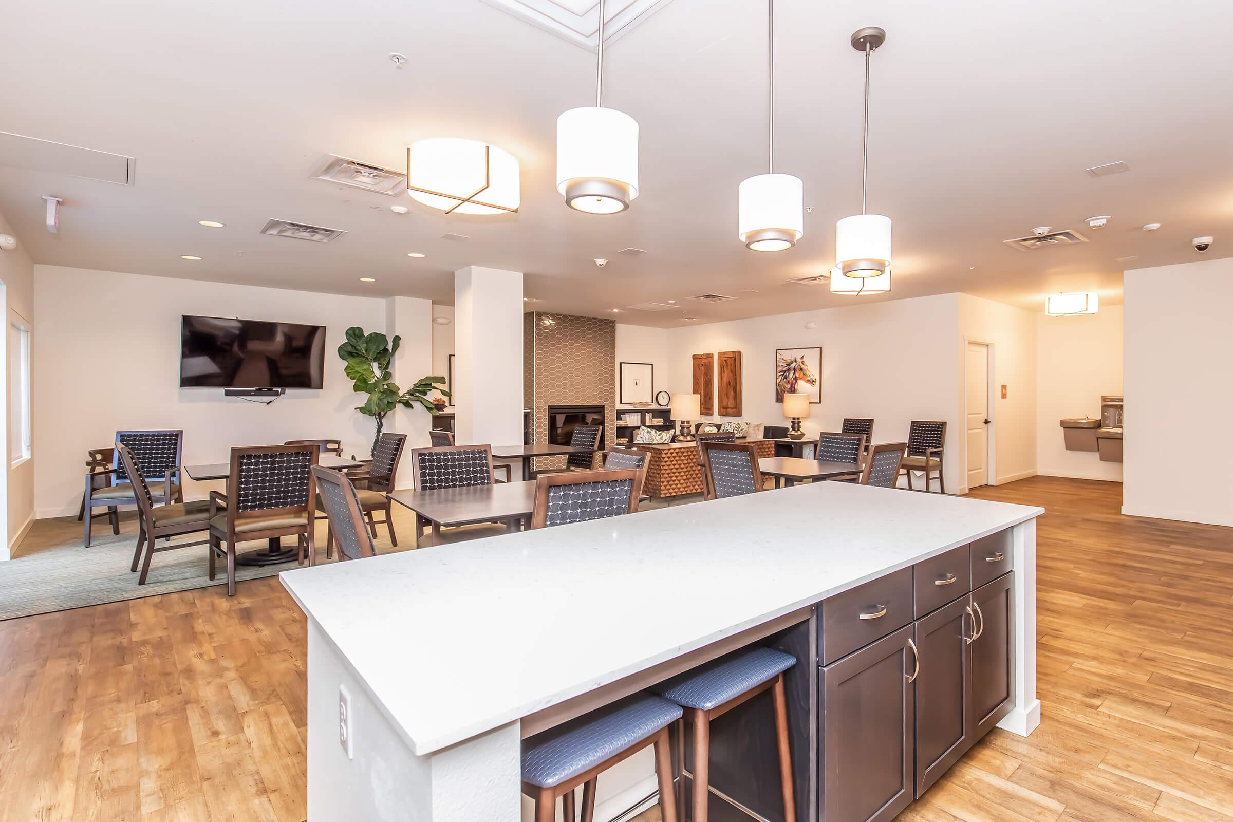 A spacious, modern common area featuring a kitchen island with bar stools, a dining space with tables and chairs, and a television on the wall. Large windows allow natural light in, and stylish decor adds a welcoming touch. The flooring is wooden, enhancing the warm ambiance.
