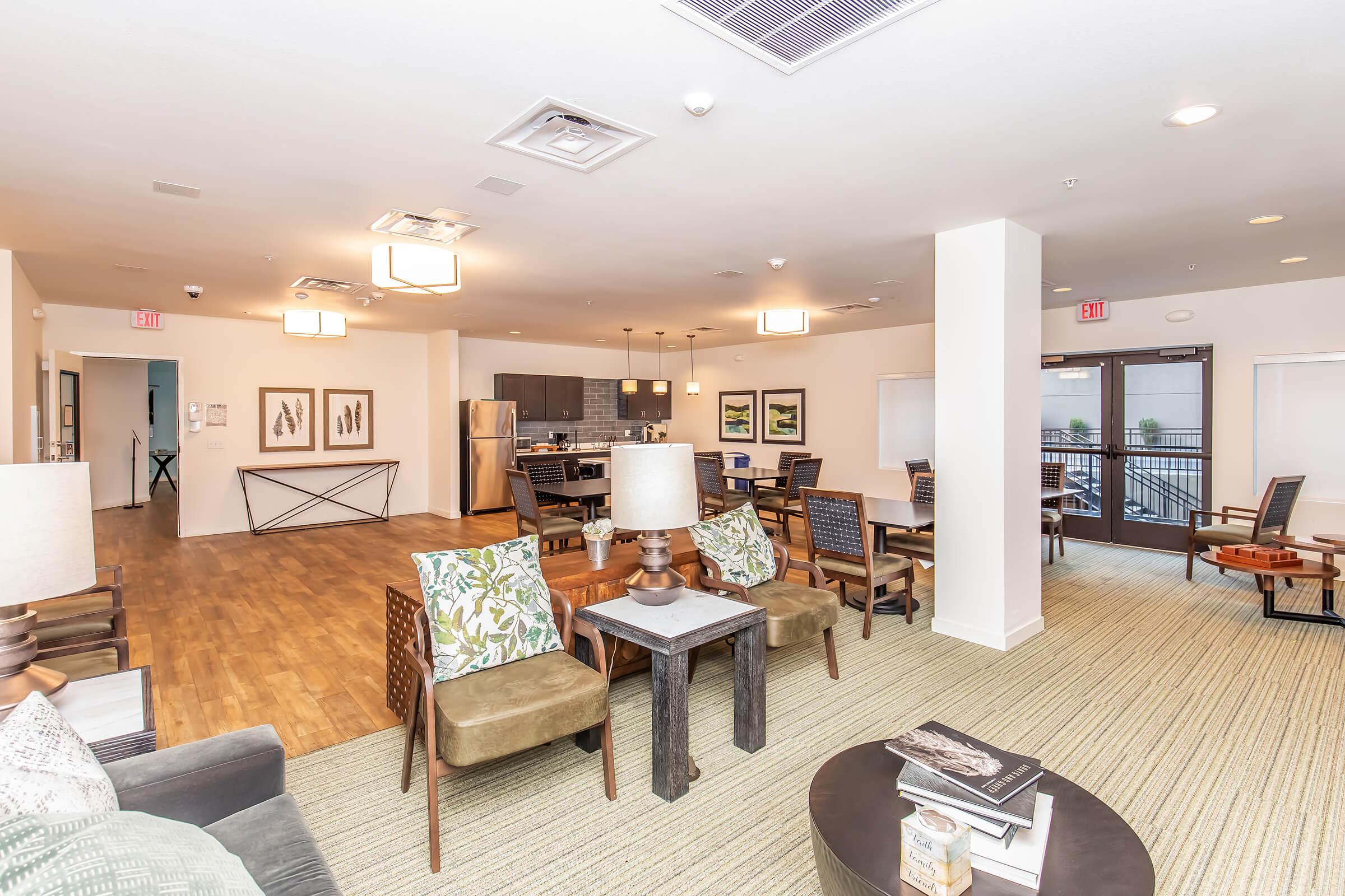 Interior view of a modern common area featuring stylish seating arrangements with patterned cushions, wooden floors, and contemporary lighting. There are dining tables and chairs in the background, alongside a kitchen area with dark cabinetry. Large windows allow natural light, creating a warm and inviting atmosphere.