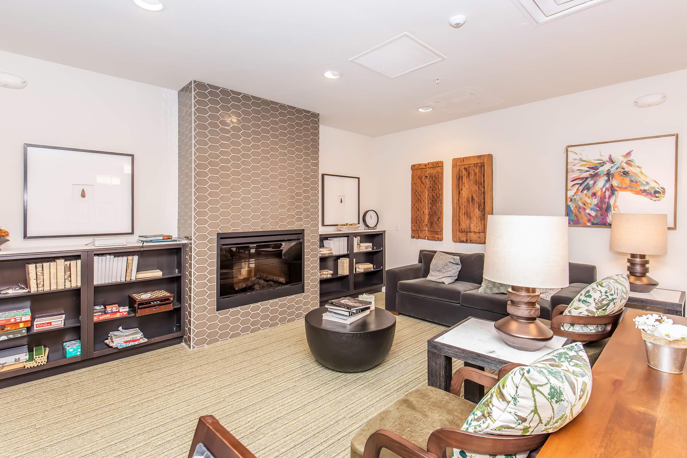 Cozy living room featuring a black sofa, decorative wooden panels on the wall, a modern fireplace with a patterned tile surround, and a stylish coffee table. Shelves filled with books and decor items are present, along with two lamps on the side tables, creating a warm and inviting atmosphere.