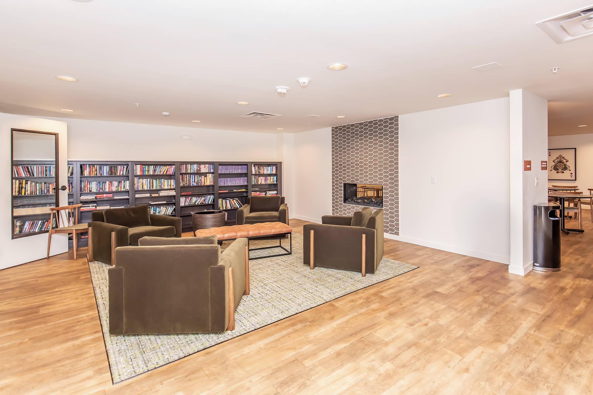 A cozy lounge space featuring comfortable seating arranged around a central coffee table, with bookshelves filled with books on one side. A decorative fireplace adds warmth to the atmosphere, while the flooring is light wooden, creating a welcoming reading and relaxation area.