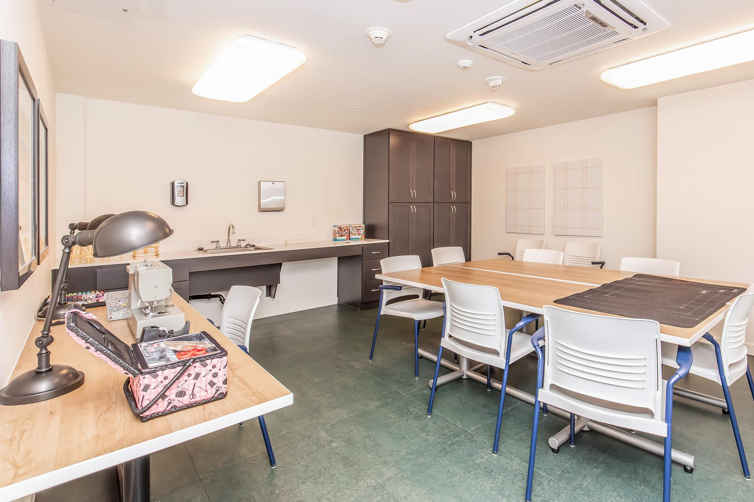 A modern meeting room featuring a large wooden table surrounded by white chairs, a kitchenette in the background, and a workspace with a lamp and supplies. The room has overhead lighting and a clean, minimalistic design, with green flooring and light-colored walls.